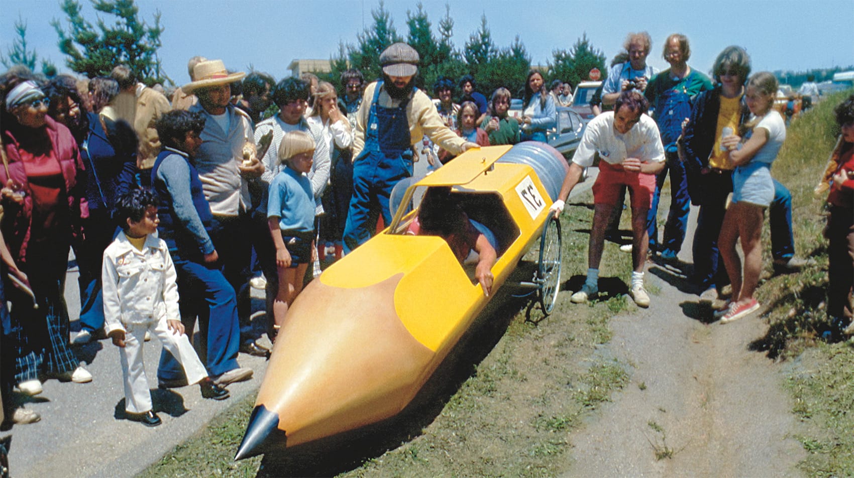 people crowd around a derby car shaped like a pencil