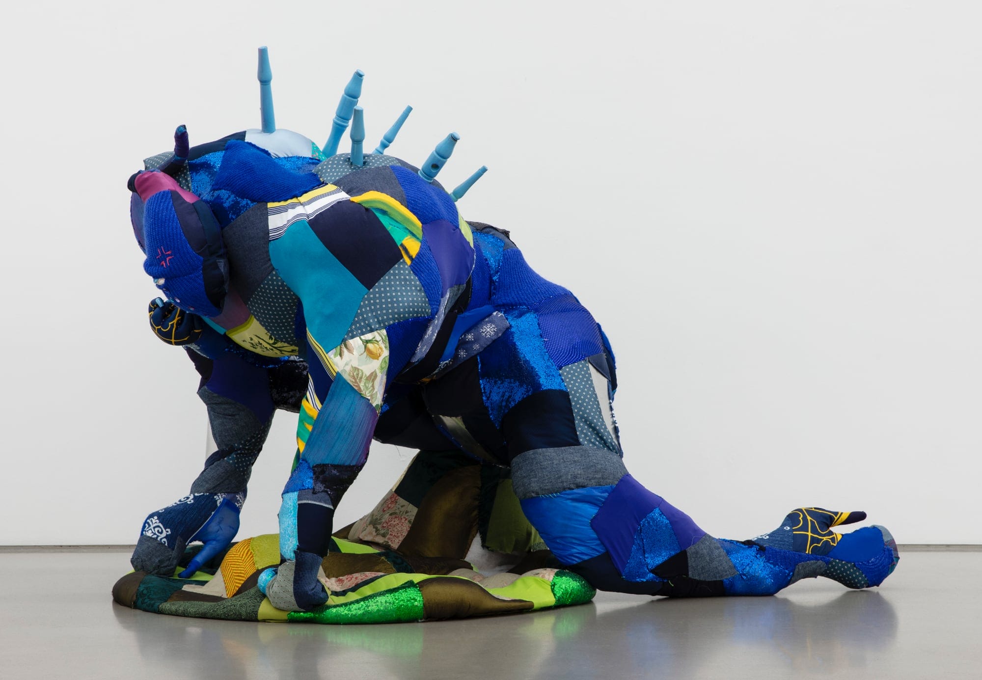 A fabric sculpture of a figure who appears crouched over, hands on the floor with spikes of blue wood emerging from its back