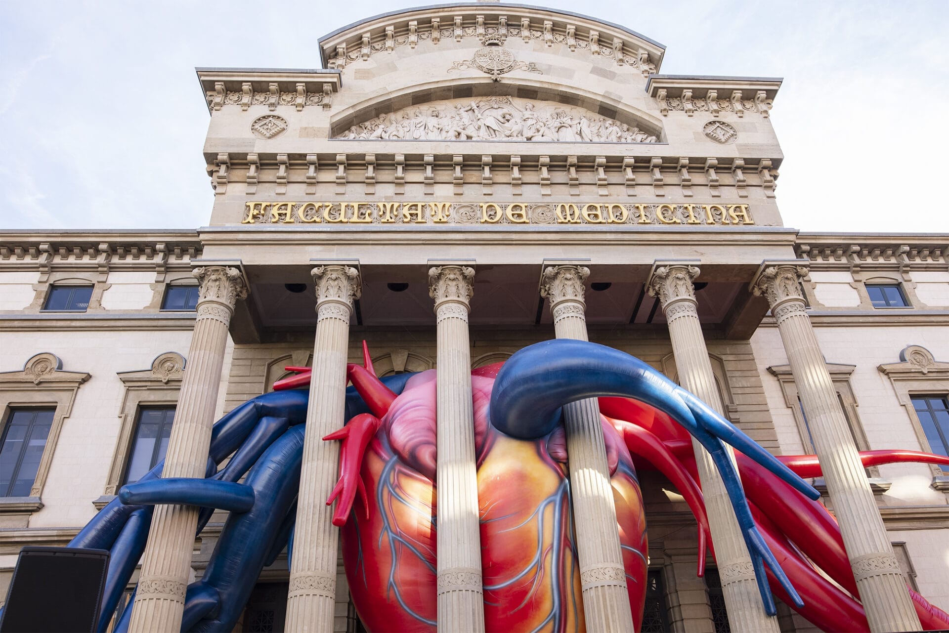 A photo of a university building front entrance with an inflatable sculpture of a realistic human heart wedged in behind its colonnade. People stand in the foreground.