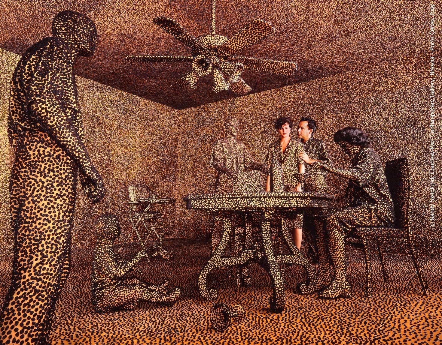 A fine art photograph featuring two people in a tableau of a living room with a table, ceiling fan, and chairs. A young child sits on the ground. Everyone and everything is covered in fuzzy black dots.