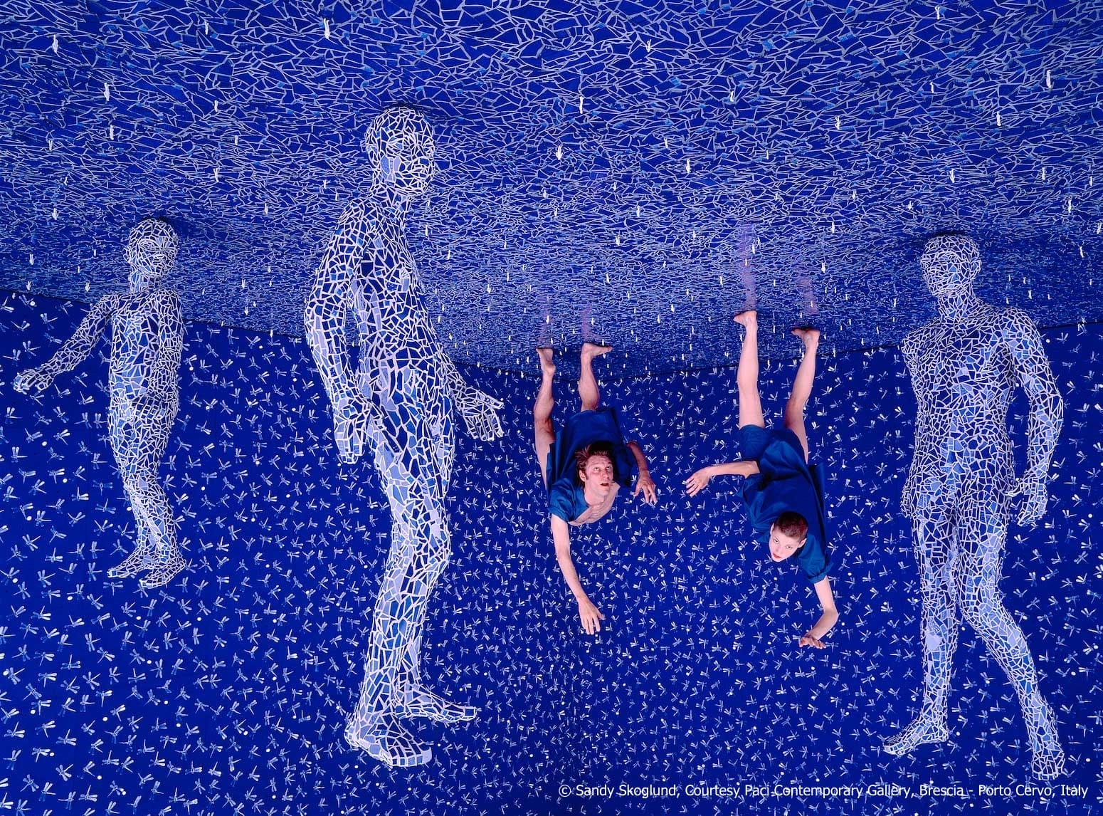 A fine art photograph of a surreal blue interior. Two figures stand on the ceiling and appear to be floating or falling, and other figures made from mosaic glass appear to be hanging from the ceiling, too.