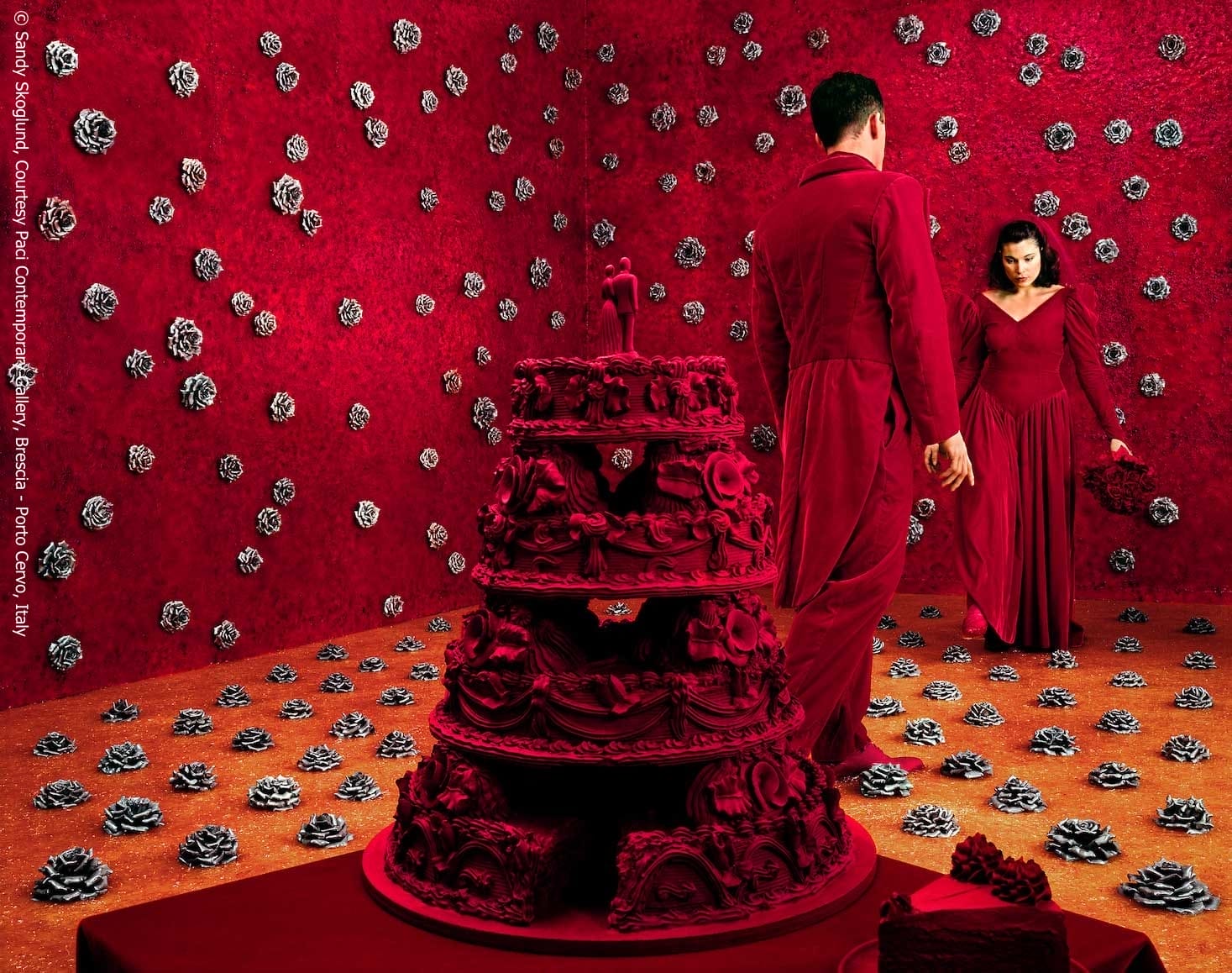 A fine art photograph of a surreal scene in which two people appear to be dancing, both wearing red velvet outfits. In the center is a large red velvet wedding cake.