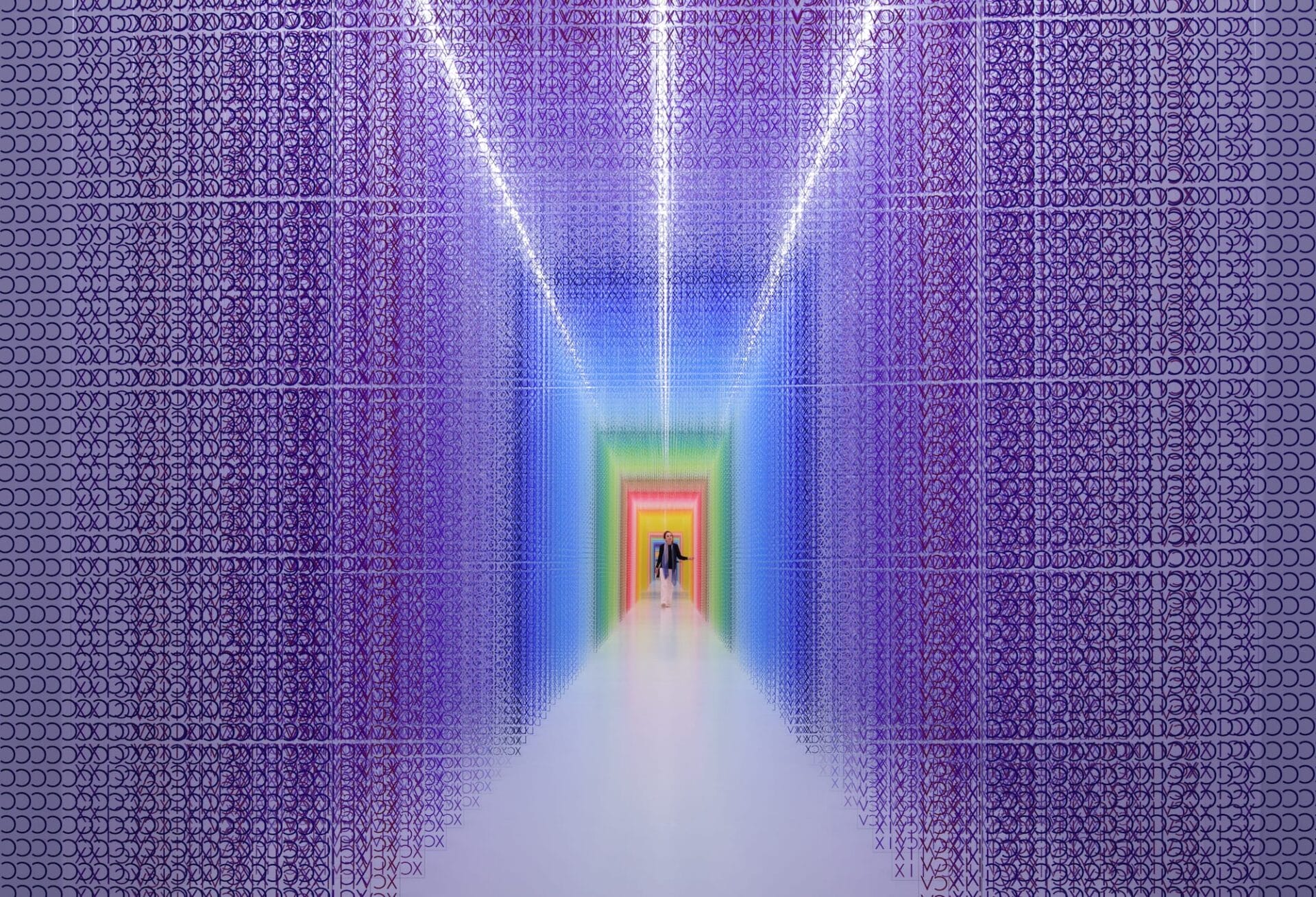 A figure walks through a large-scale installation made from thousands of brightly colored Roman numerals in a corridor that gradually changes color.
