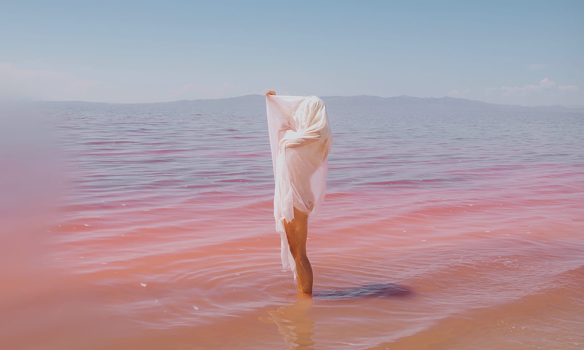 A photograph depicting a figure covering themselves in a wet-looking white cloth or garment standing ankle-deep in a body of water. The water reflects from blue to pink the closer it is to the foreground.