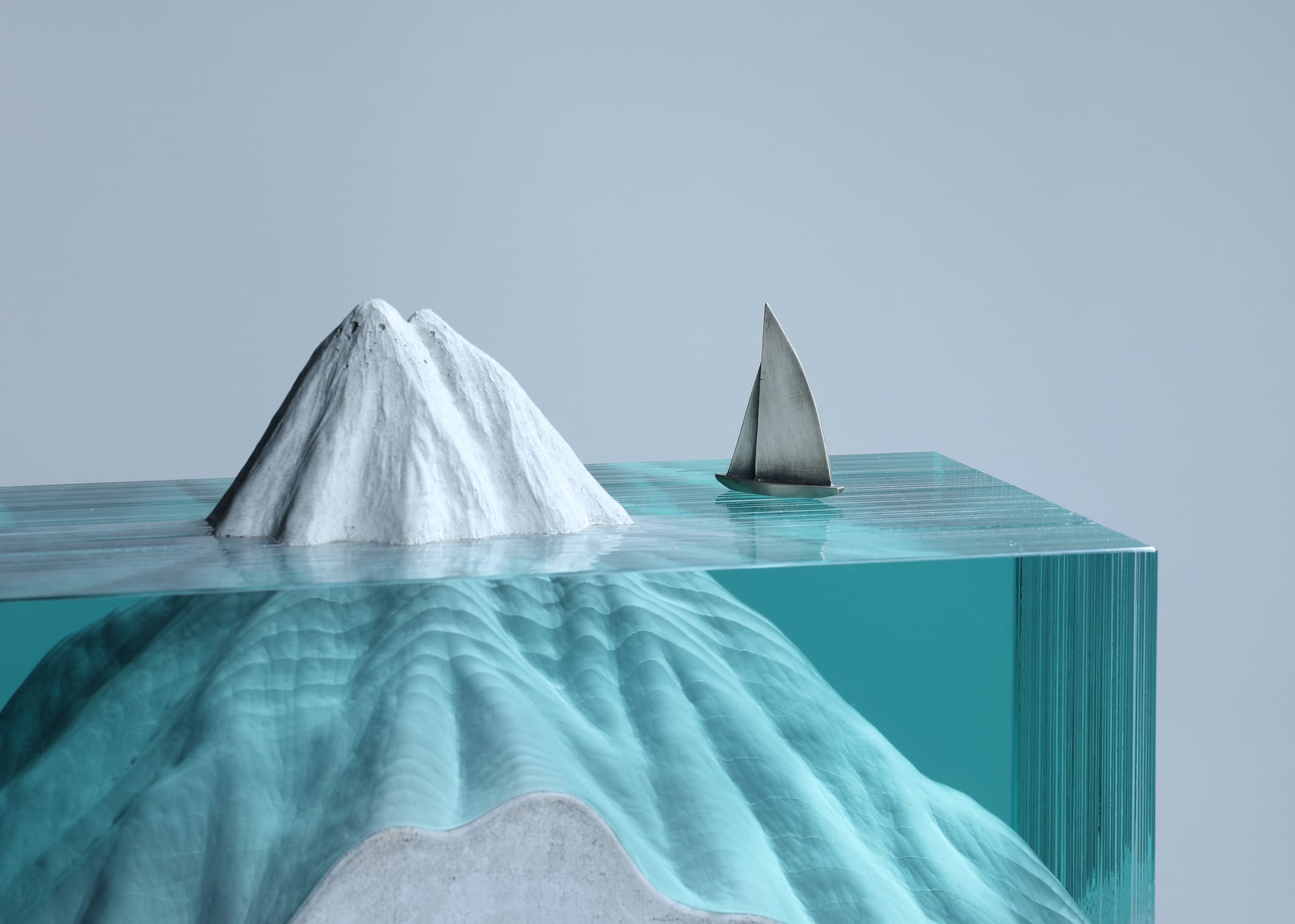 A sculpture made of concrete, steel, and layers of glass that depicts a mountain appearing on the surface of some water, with a sailboat nearby.