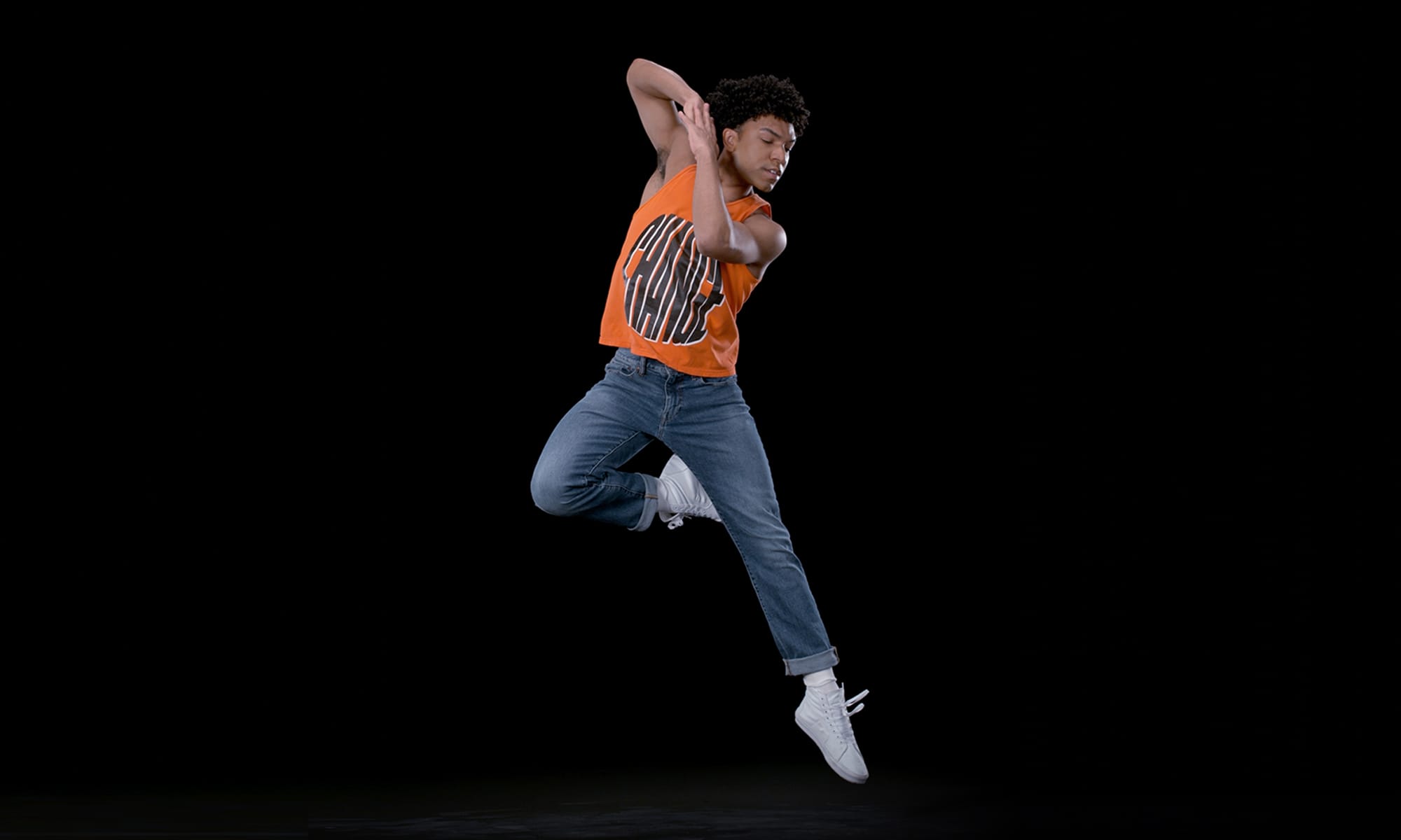 A ballet dancer in blue jeans, an orange t-shirt, and white sneakers leaps into the air.