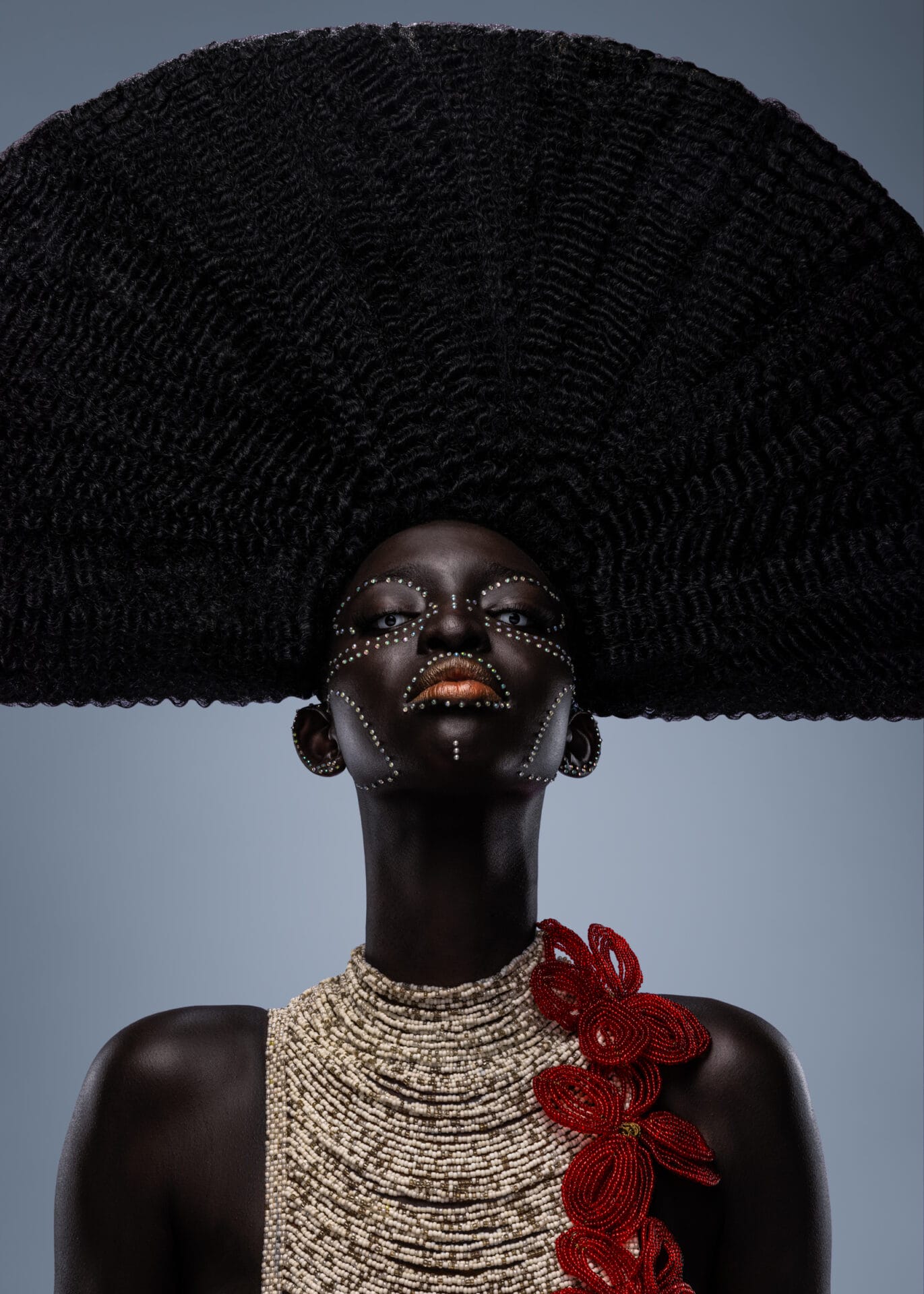 A portrait of an African woman donning traditional beadwork, styled hair, and ornate garments. She focuses her eyes toward the camera lens.