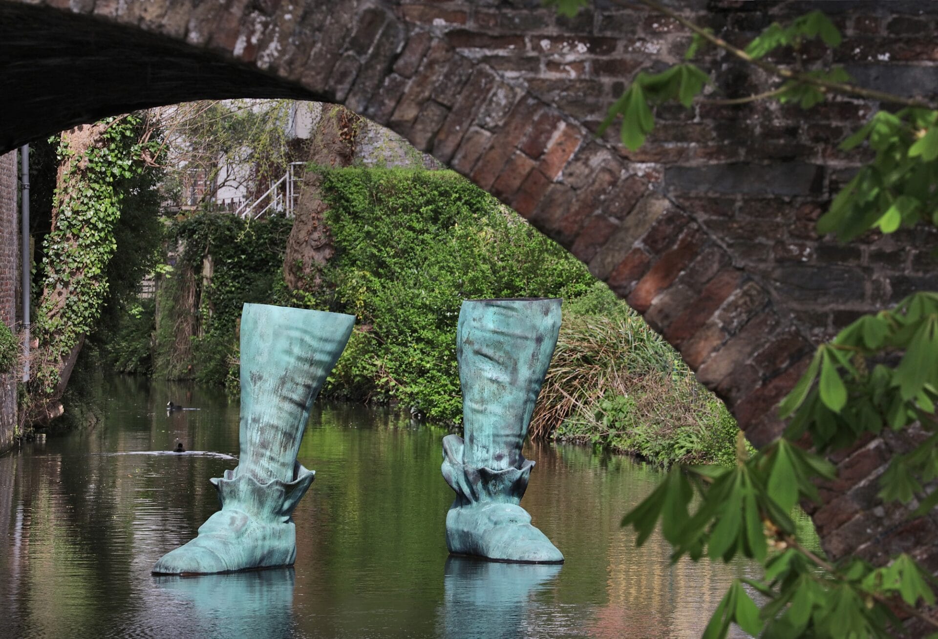 a sculpture of a pair of 18th-century style boots cast in bronze and installed in a Bruges canal