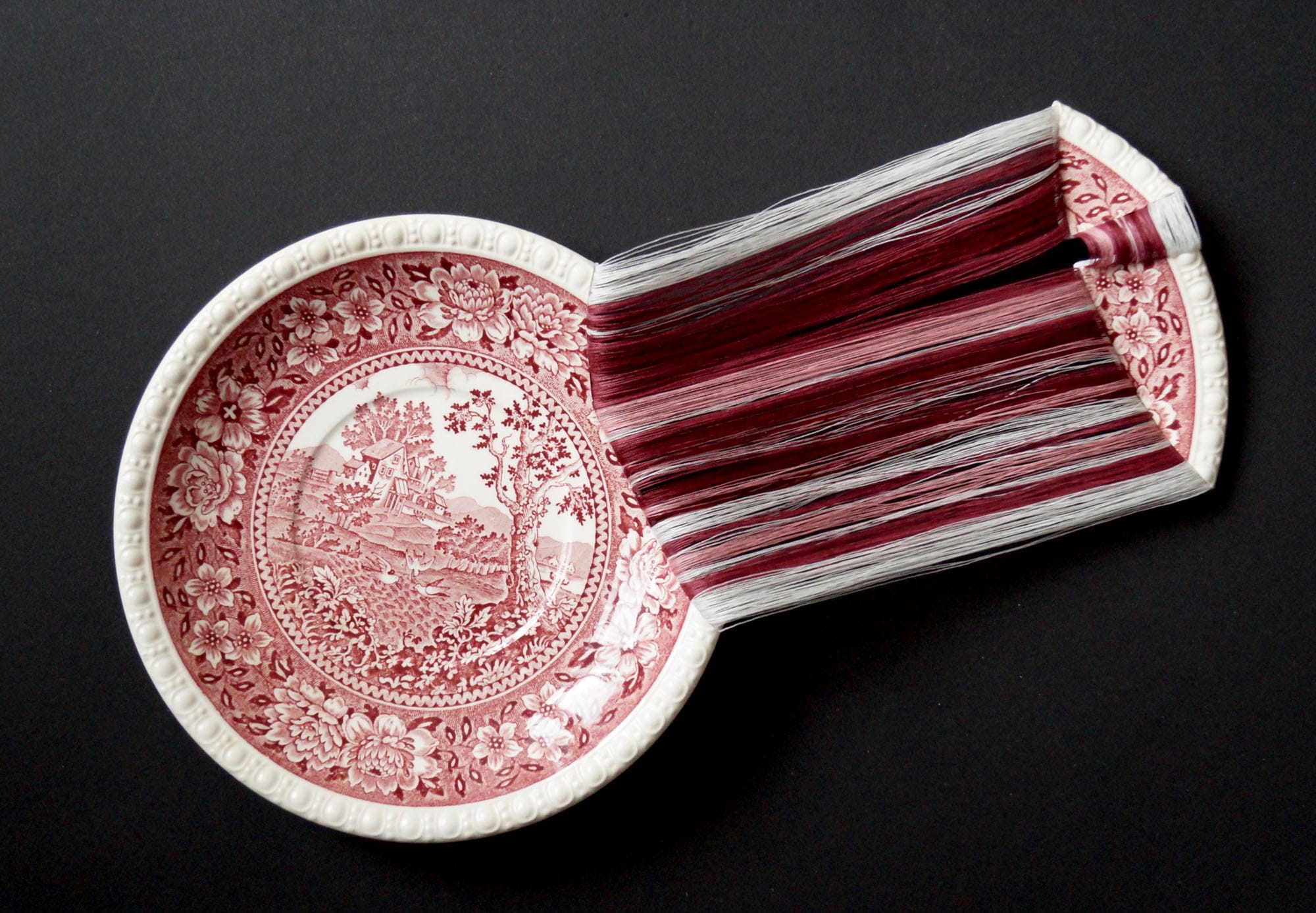 Hundreds of filigree threads hang from a broken porcelain plate in hues of red and white.