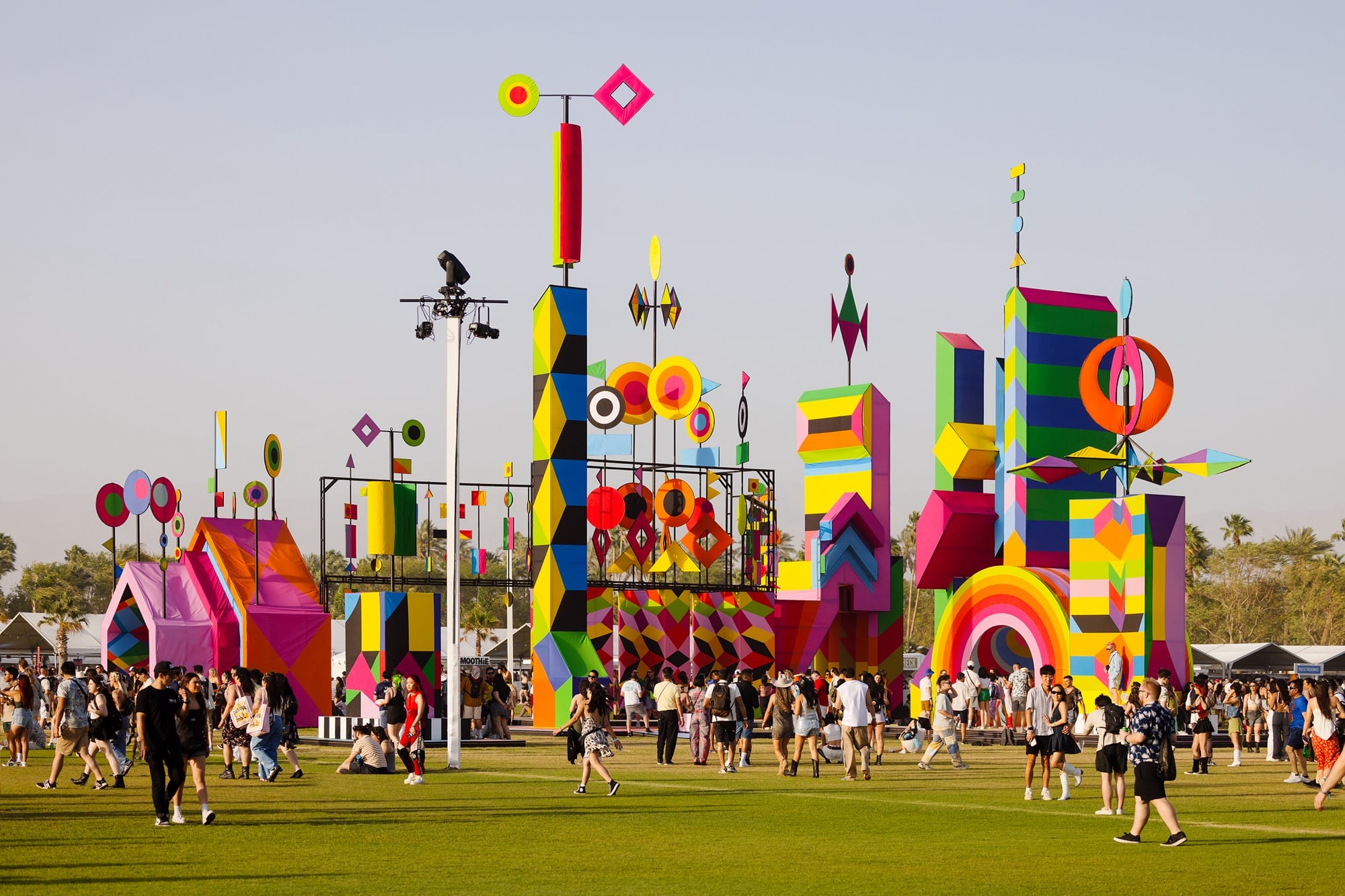 a large-scale, colorful public installation at Coachella music festival showing lots of geometrically-patterned columns and kinetic details that spin in the wind as people gather below on a colorful floor