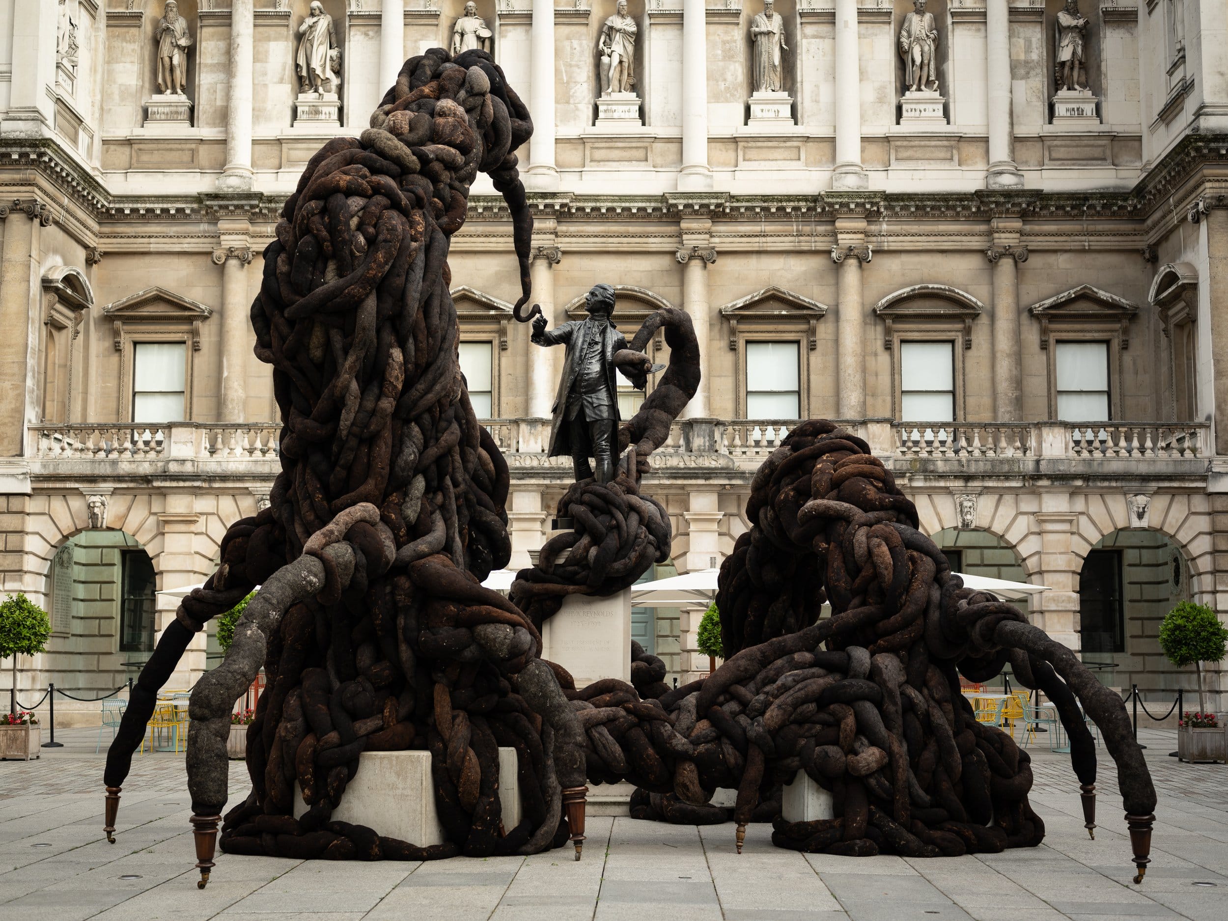 a large-scale sculpture in the courtyard of the Royal Academy in London showing a fantastical creature made of horsehair and netting, rising up to meet a bronze statue of Sir Joshua Reynolds