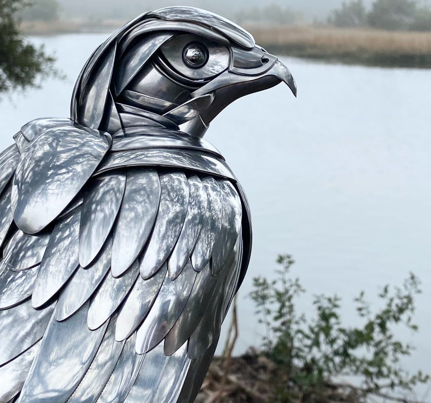 a metal bird made from repurposed silverware perches in the trees, a body of water rests in the background