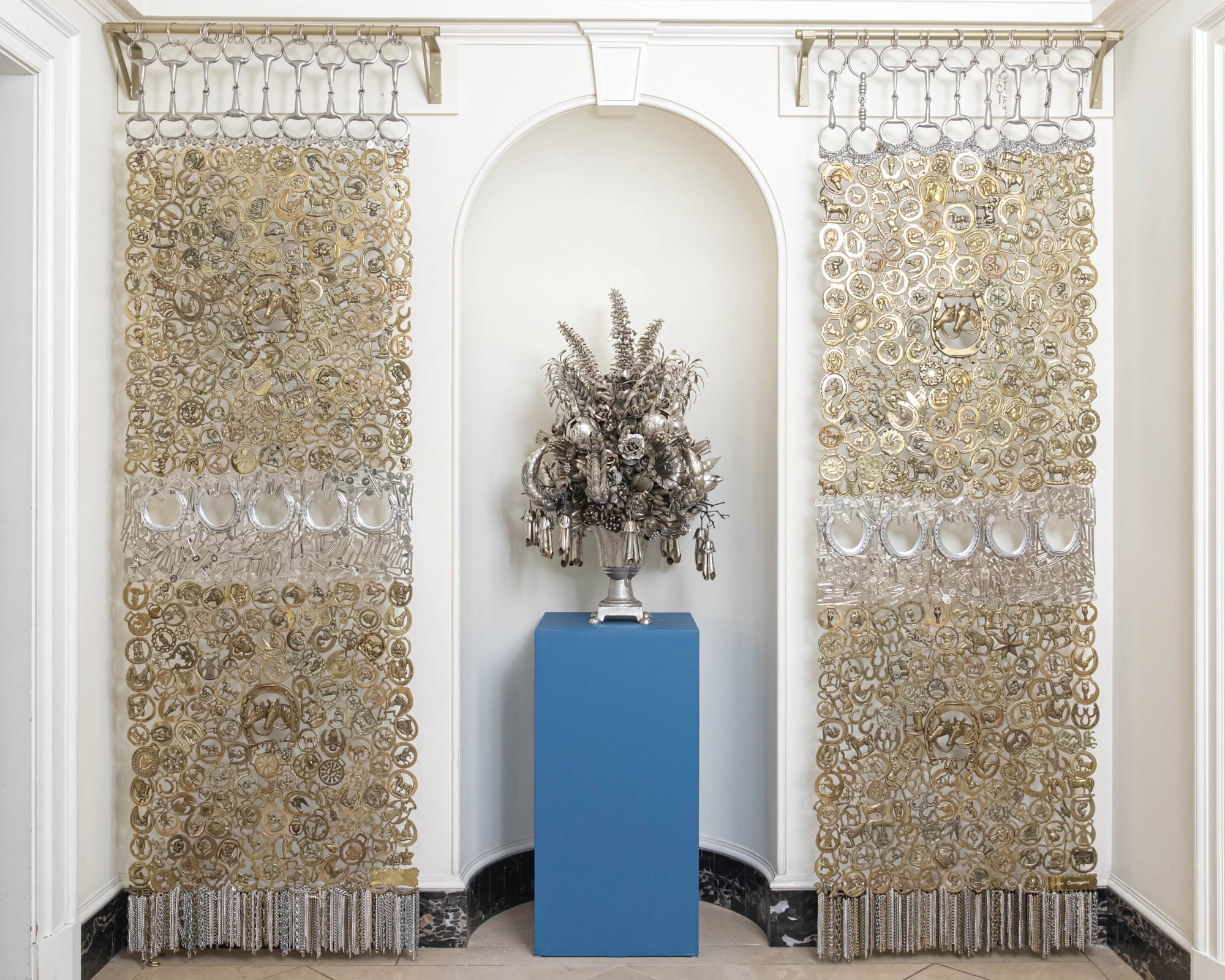 an installation view of metal mesh screens and a large floral arrangement in the center on a blue pedestal, all made out of found metals like cutlery, horseshoes, and decorative metals