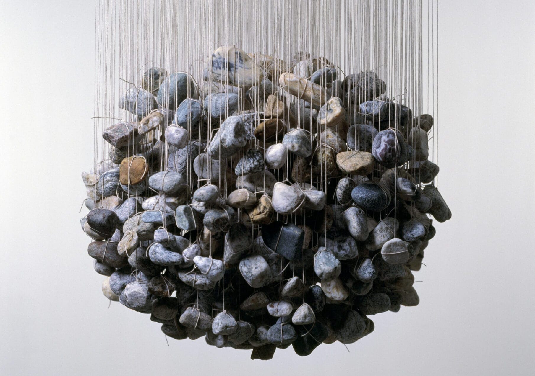 a detail image in a gallery space, many river stones are suspended from the ceiling with string. the stones hang at different heights, forming a spherical shape