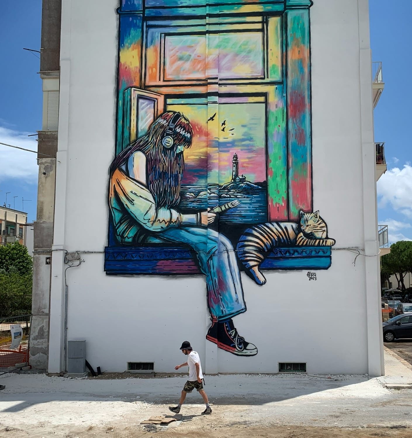 on the side of a building, a mural depicts a girl sitting on a windowsill with a book as a cat lounges next to her.