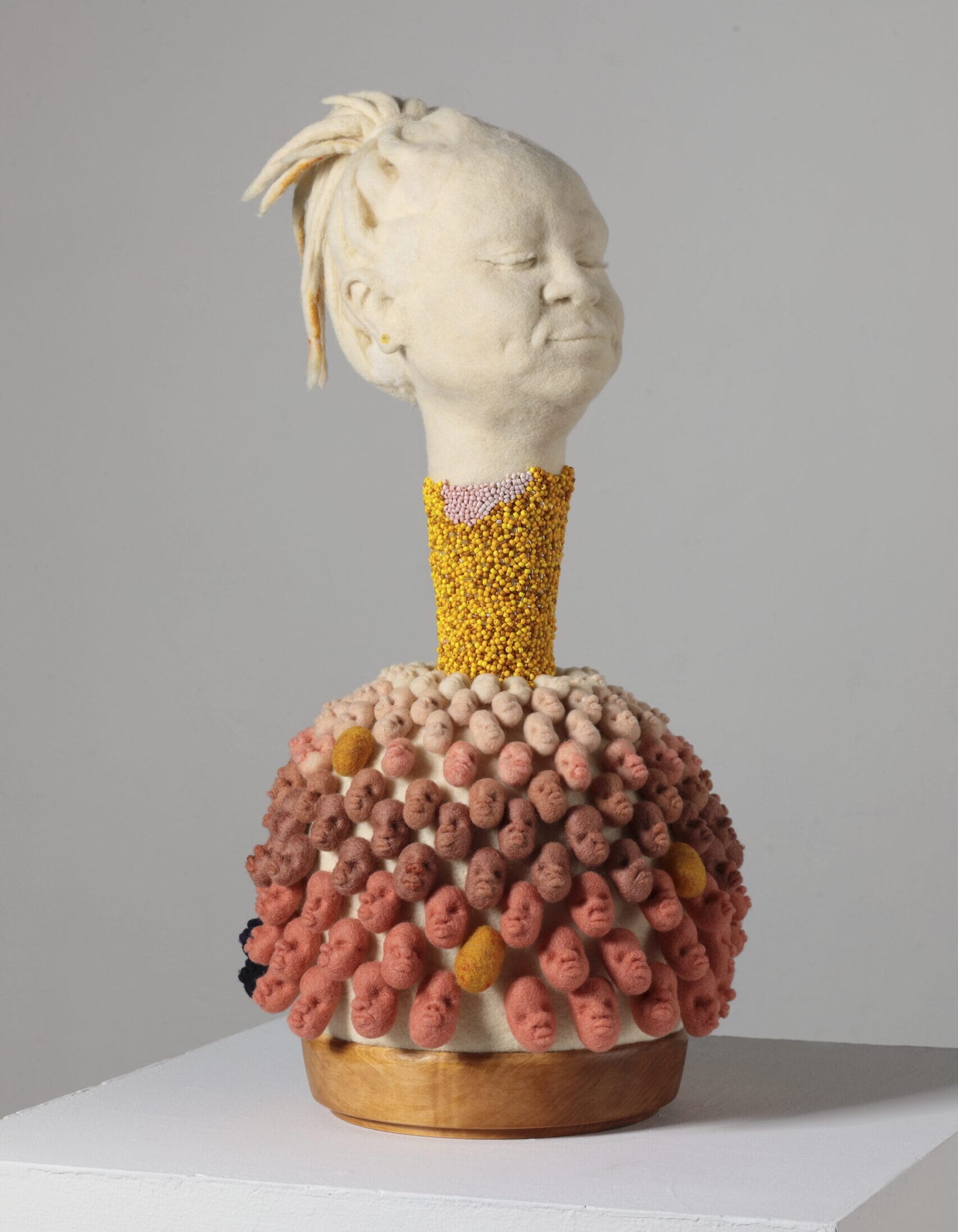 a sculptural bust made from felted wool showing a Black woman's head on a beaded pillar with numerous faces at the base in a gradient of skin tones