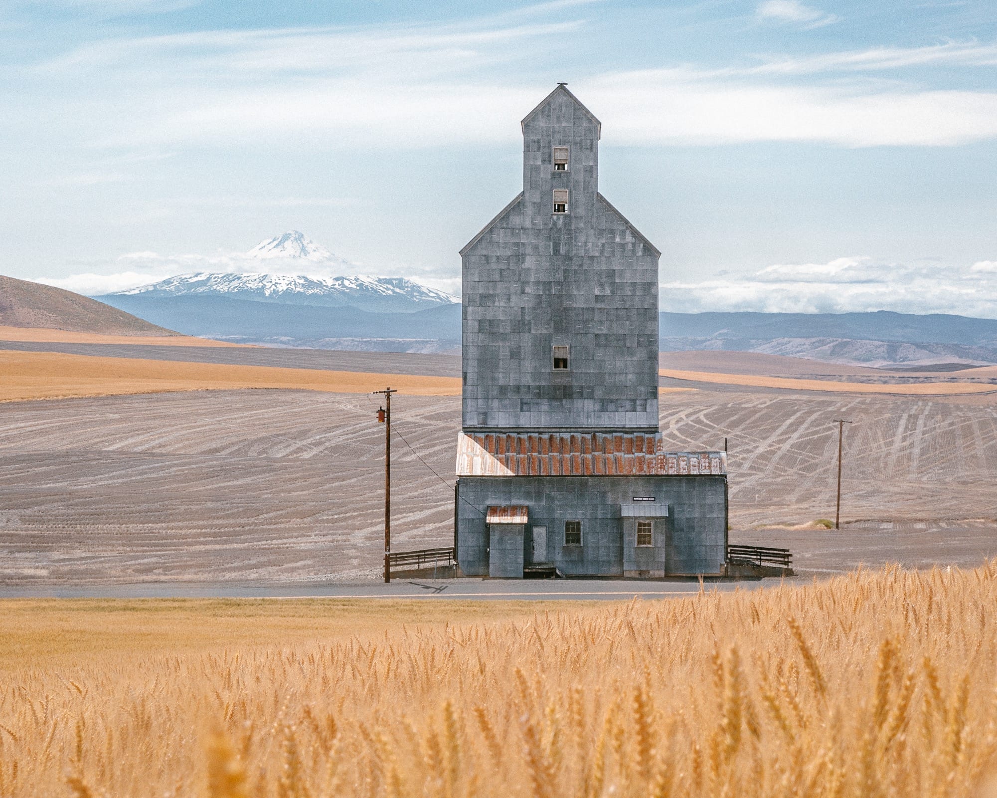 a photograph of wheat fields and a grain elevator in the foreground of some snow-capped mountains in the distance
