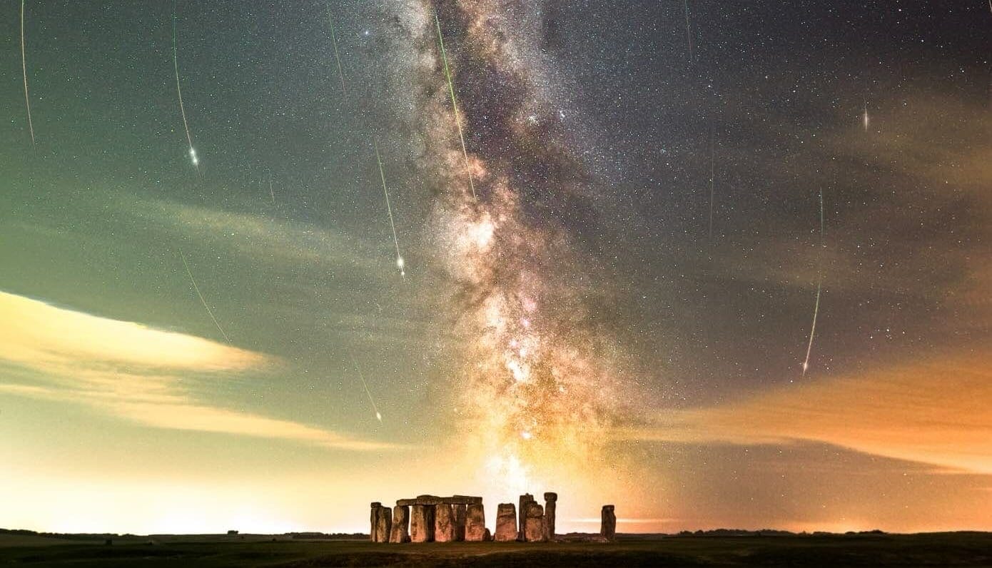meteors streak down the sky to frame the milky way, while stonehenge sits at the base