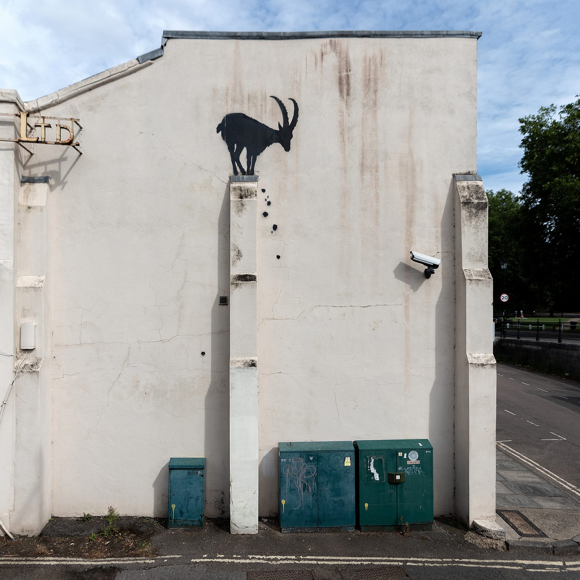 a mural of a small goat on the side of a building, standing on top of a buttress with pebbles falling at its feet