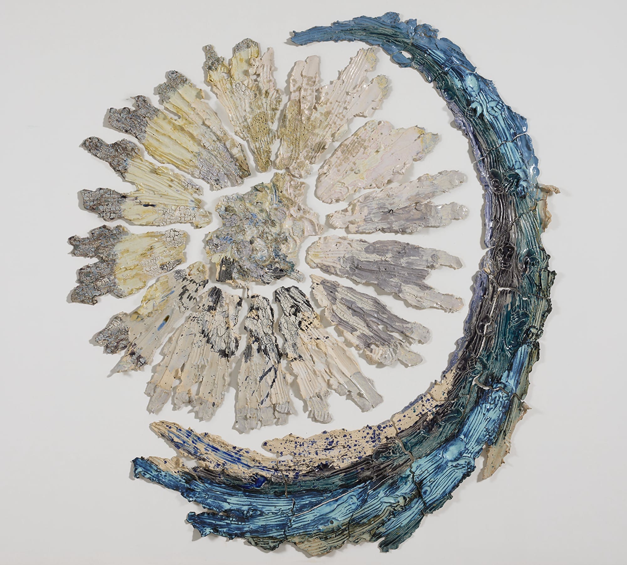 a round beige and white wall sculpture that shows pieces of ceramic with lines evocative of stretching radiating from a central point. a curved blue piece cradles the right and bottom sides
