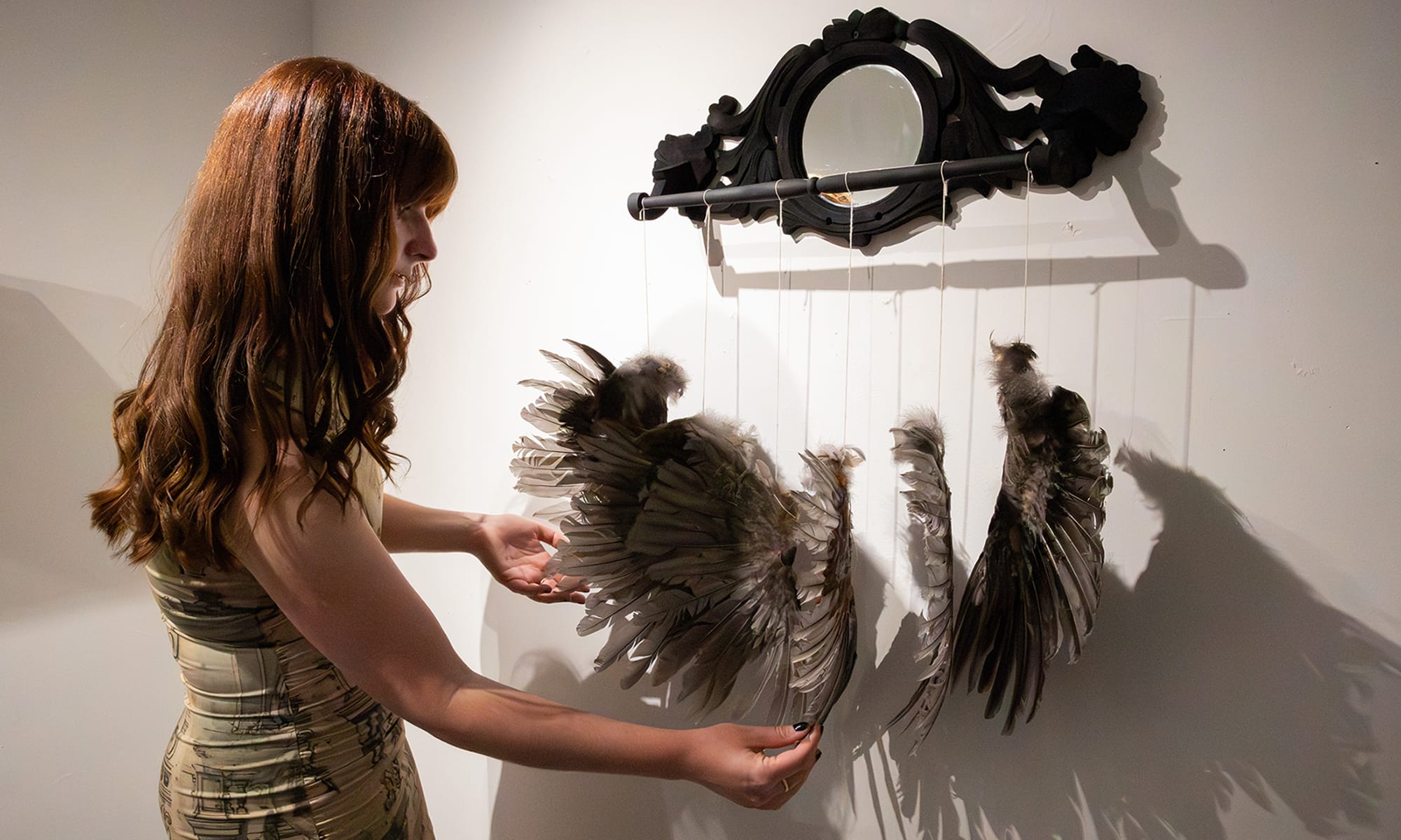 a white woman with red hair adjusts a feather sculpture that's dangling from an ornate black mirror