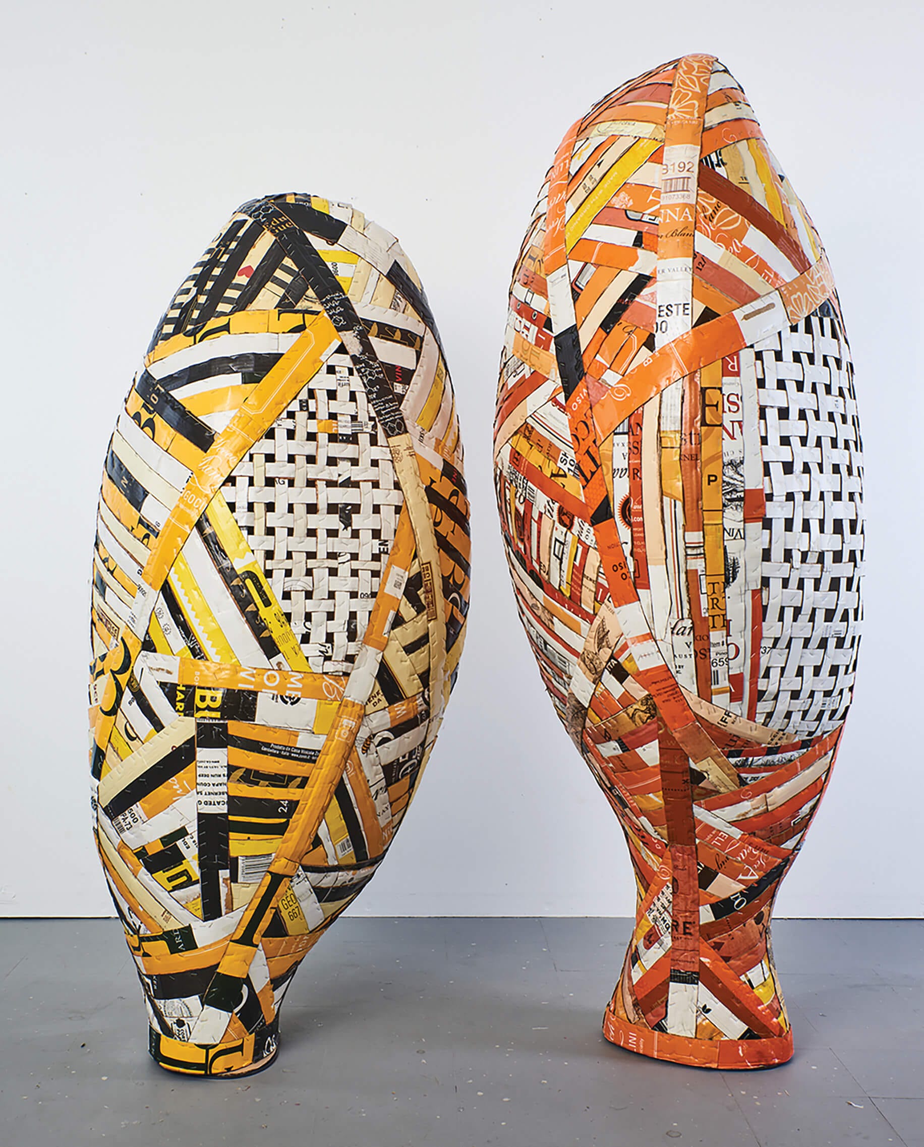two freestanding sculptures side-by-side made of woven and layered strips of colorful cardboard. the one on the left is primarily yellow, black, and white, while the one on the right is mostly orange and white