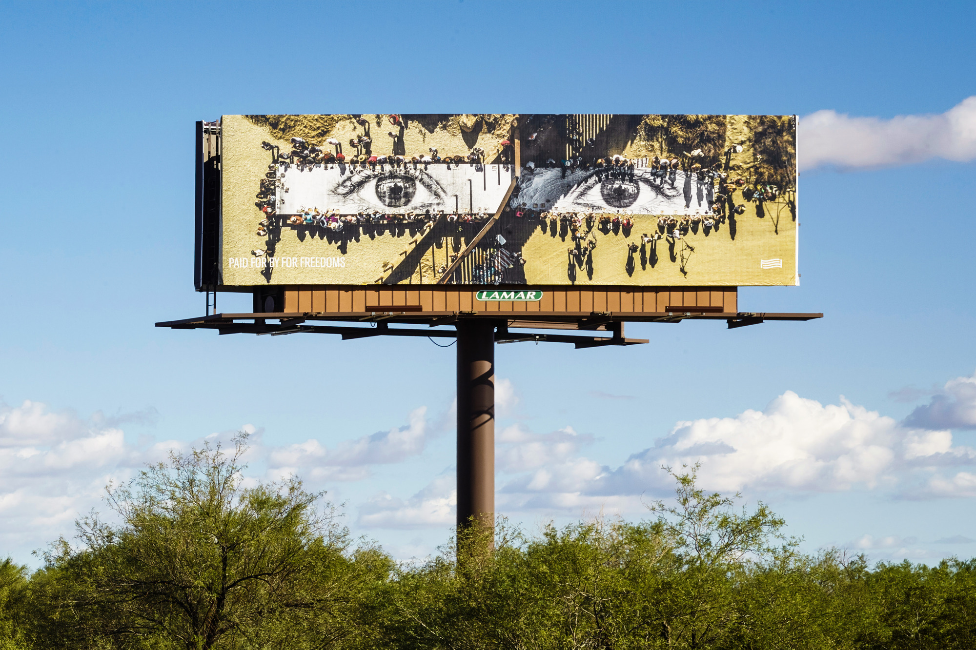 a billboard sign with an photograph of a public project involving dozens of individuals holding a large, long artwork print of two open eyes.