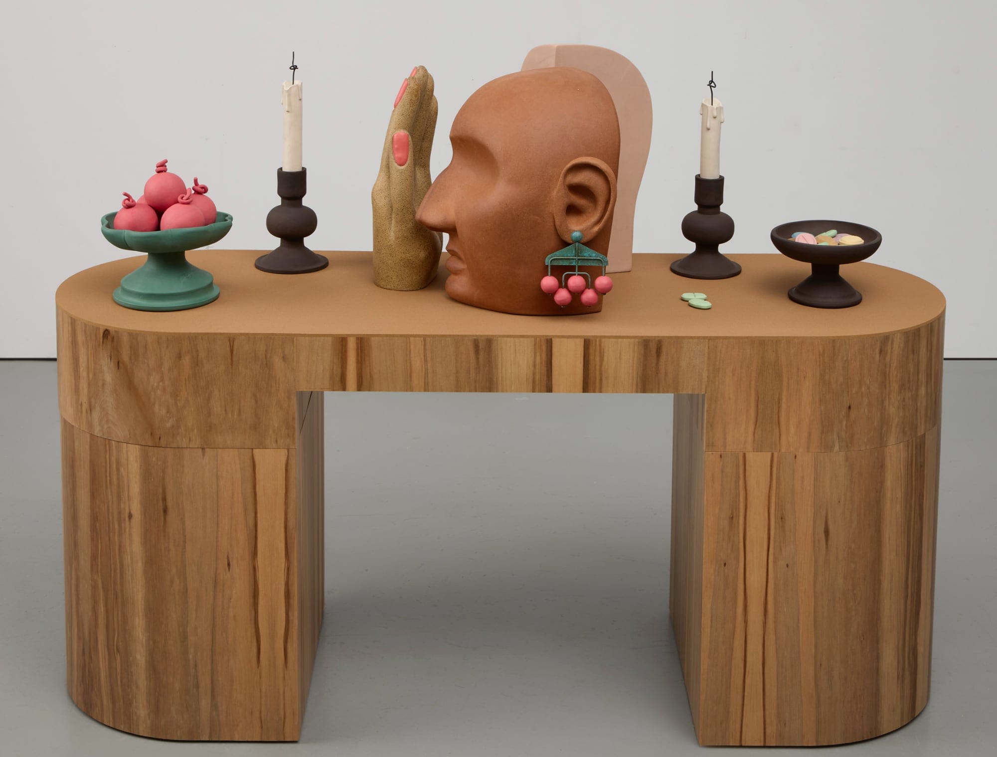 fruit in a bowl, a candle stick, a hand with pink nails, a face with pink dangling earring, another candlestick and bowl rest atop a wooden table