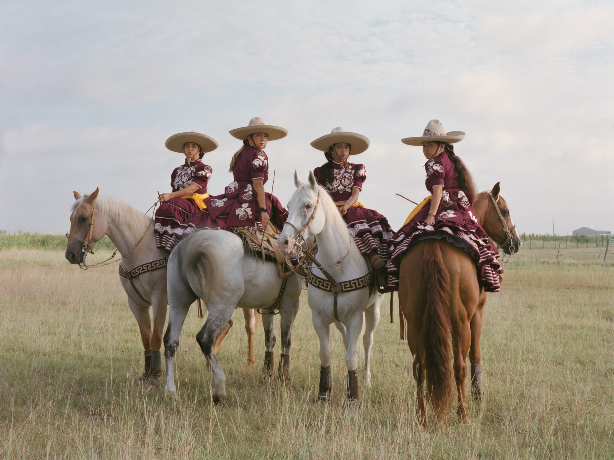 four women sit on horseback in a prairie, wearing colorful dresses and wide-brimmed hats
