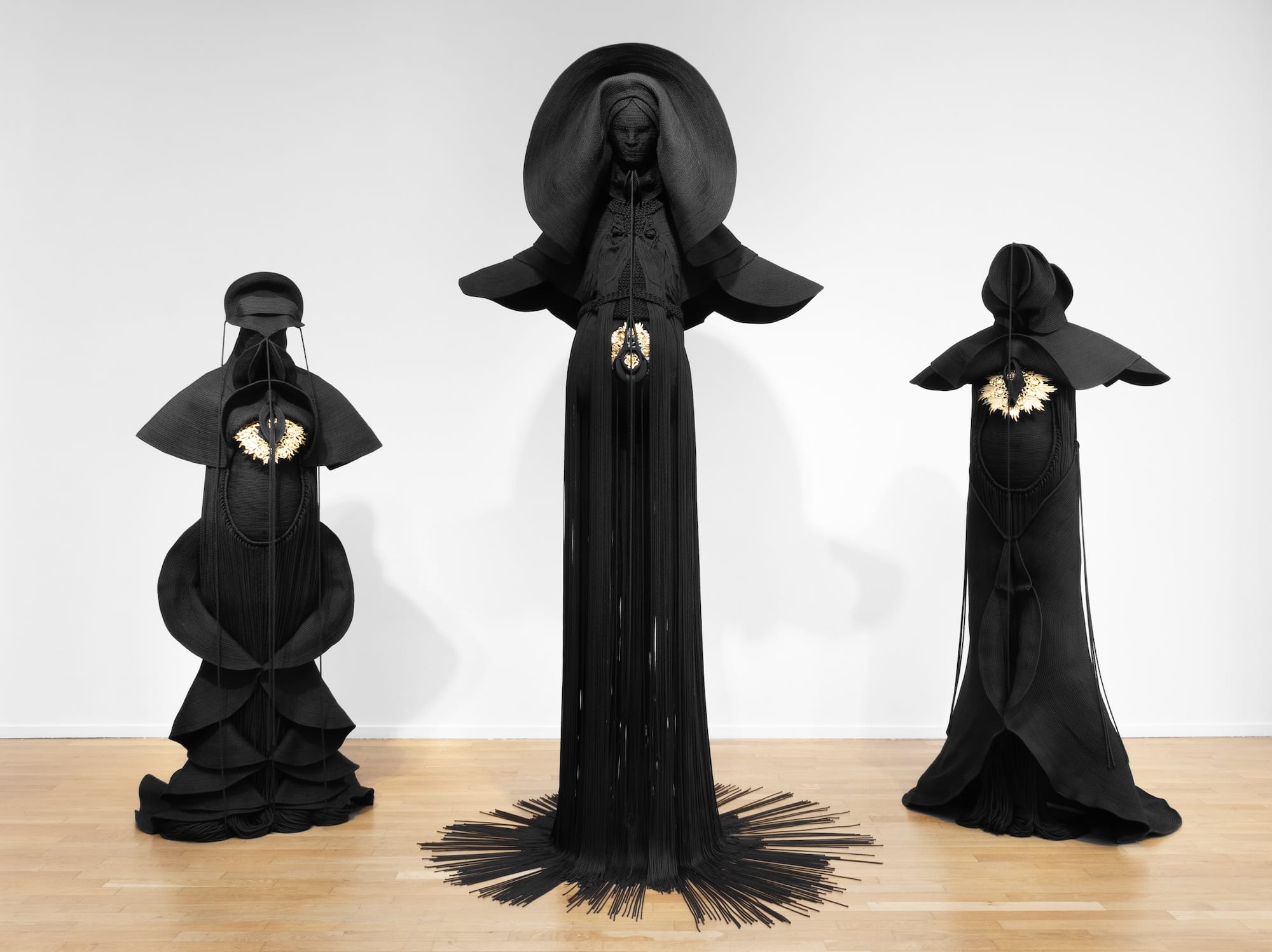 Installation view of three sculptures made from black cord, thread, and metal in the form of figures or long, dramatic garments