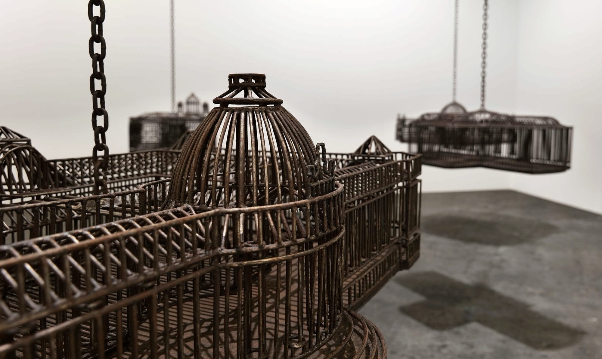 detail of an installation in a white-walled gallery space of sculptures suspended from the ceiling by chains, each one modeled after a colonial, institutional building