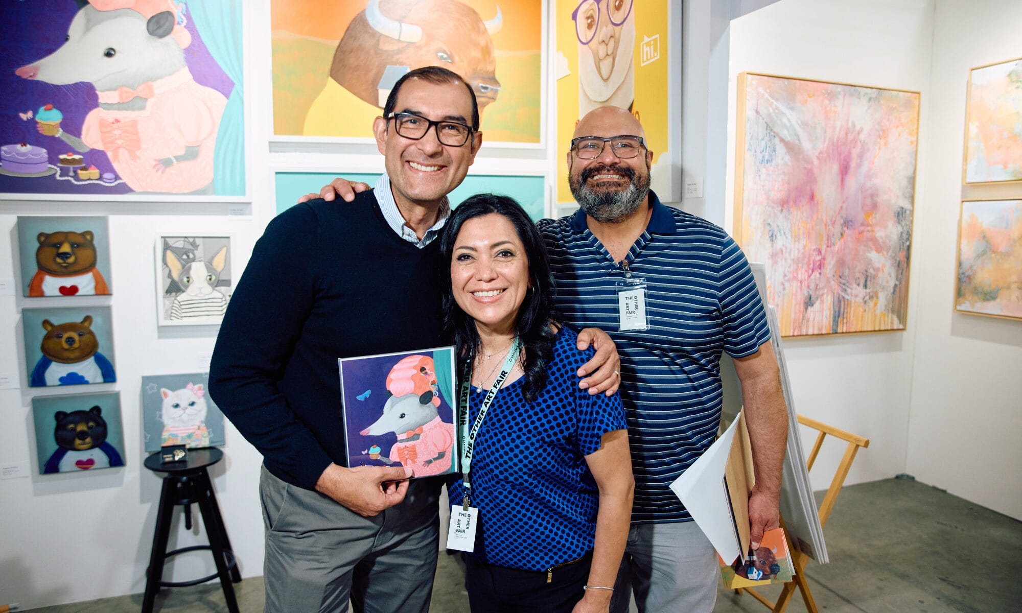 Photograph of three people standing in front of a wall of paintings.