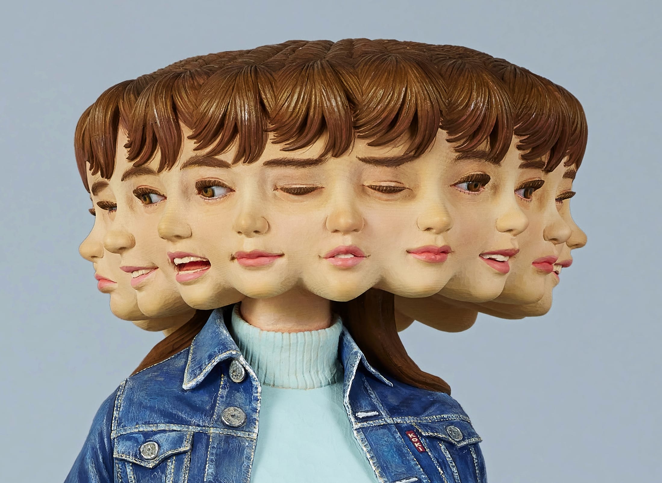 a detail of a wooden sculpture by Yoshitoshi Kanemaki of a female figure with a bright pink skirt and a denim jacket, with multiple faces and expressions