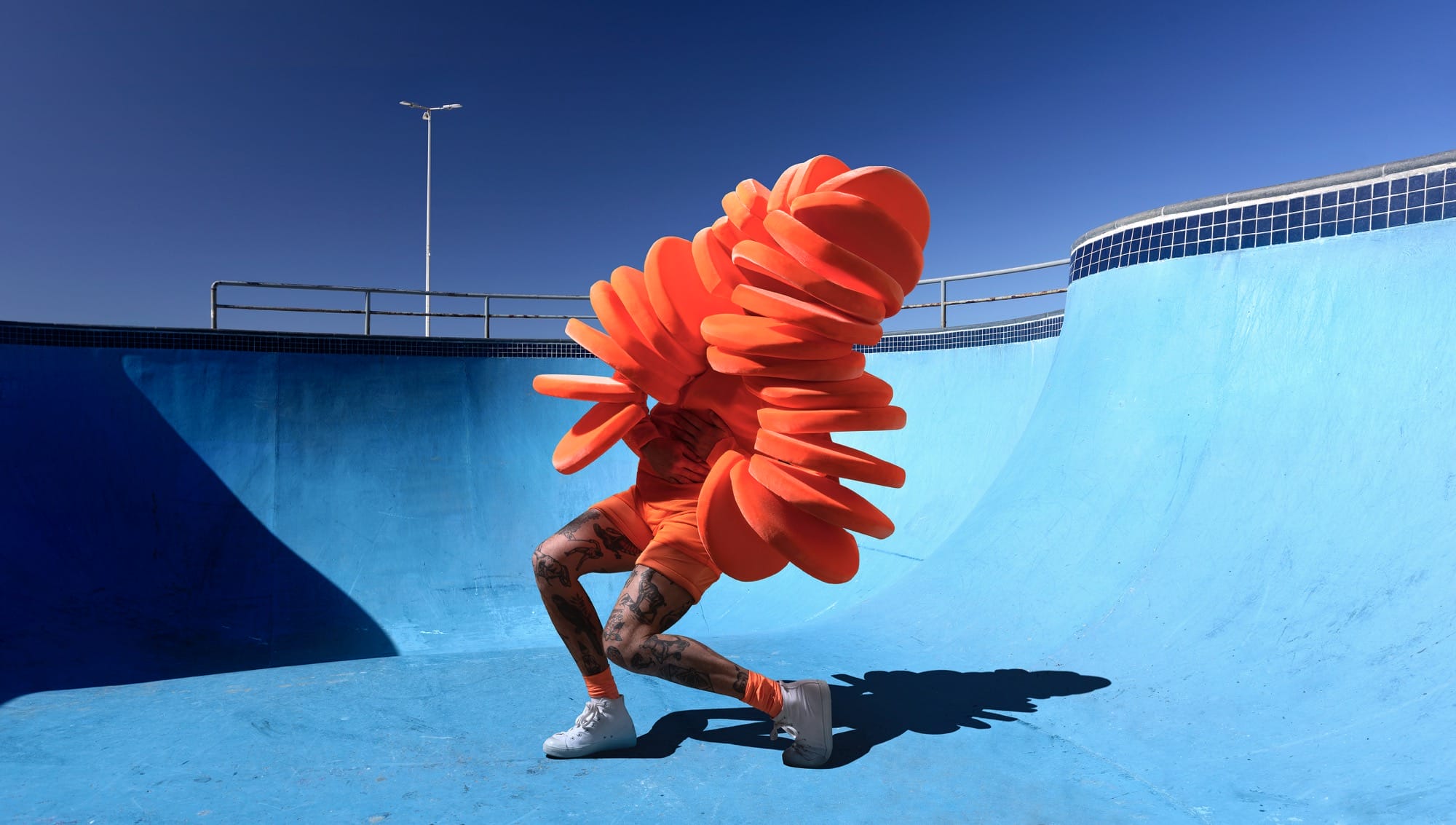 a photograph by Gerwyn Davies of a brightly colored abstract costume obscuring the top half of his body in a blue-painted skatepark