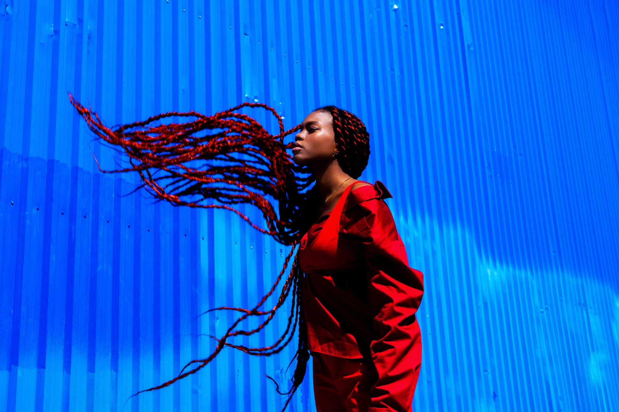 a photograph by Yannis Guibinga of a Black woman with long black-and-red braids, wearing a red outfit and standing in front of a bright blue wall