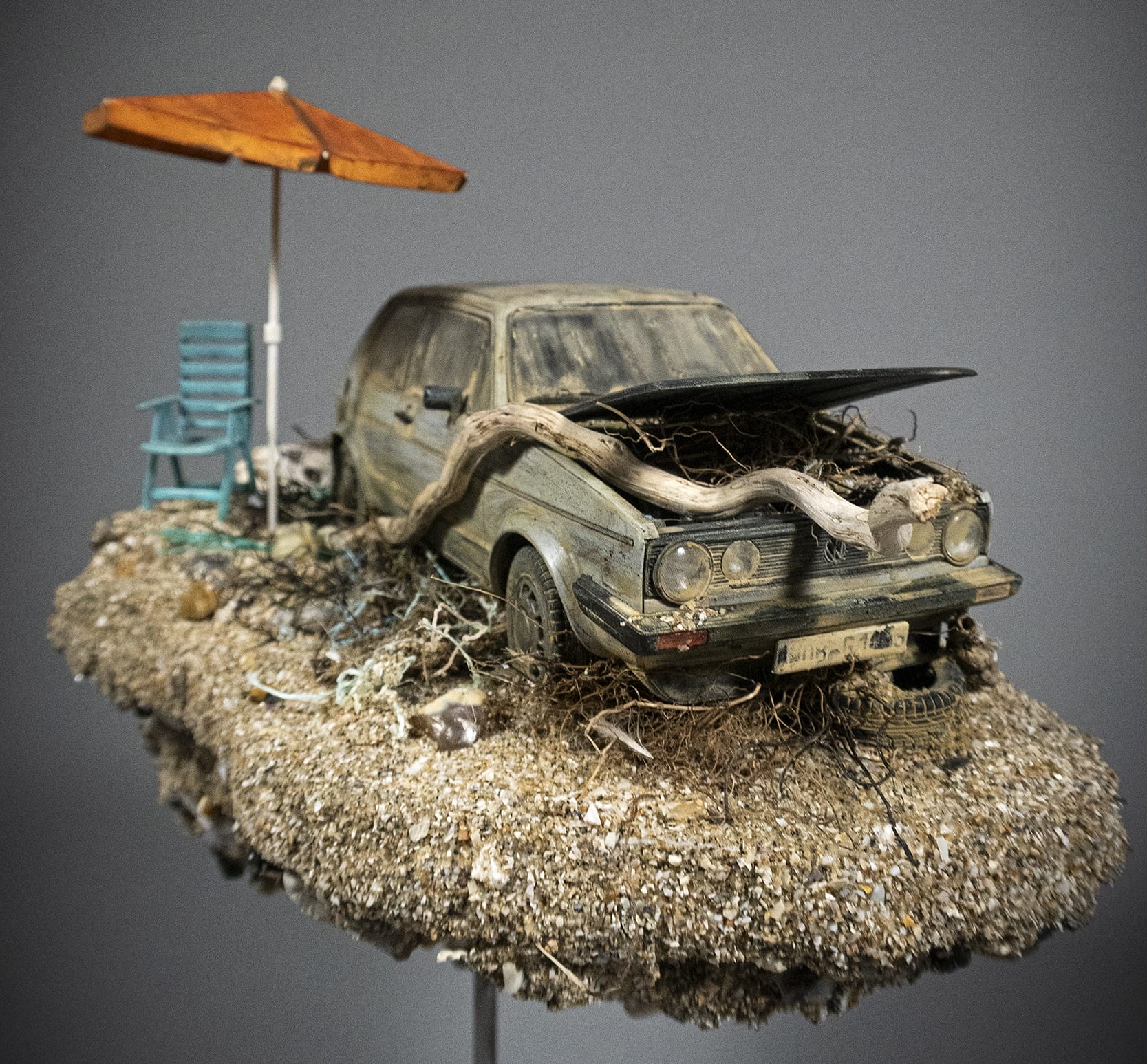 a hand-sculpted miniature by Simon Laveuve of an abandoned car with a large root growing through the hood, with a beach umbrella propped up next to it