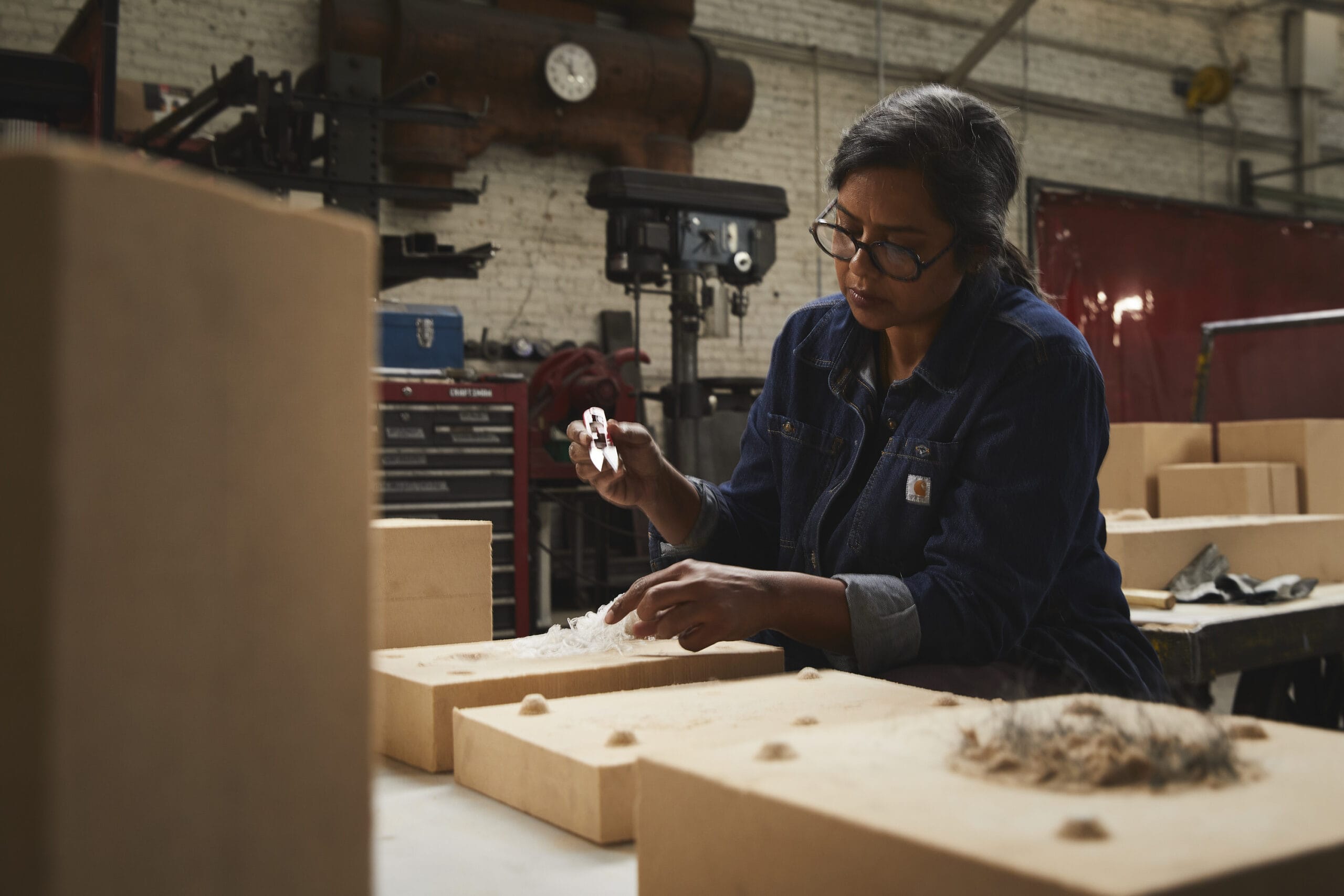 A photograph of artist Nirmal Raja working on a piece inside a warehouse. She is holding pliers on her right hand and holding the work down with the other.
