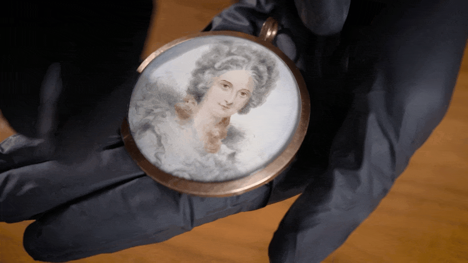 a gif of black gloved hands flipping over a locket with a portrait on one side and a lock of hair on the other