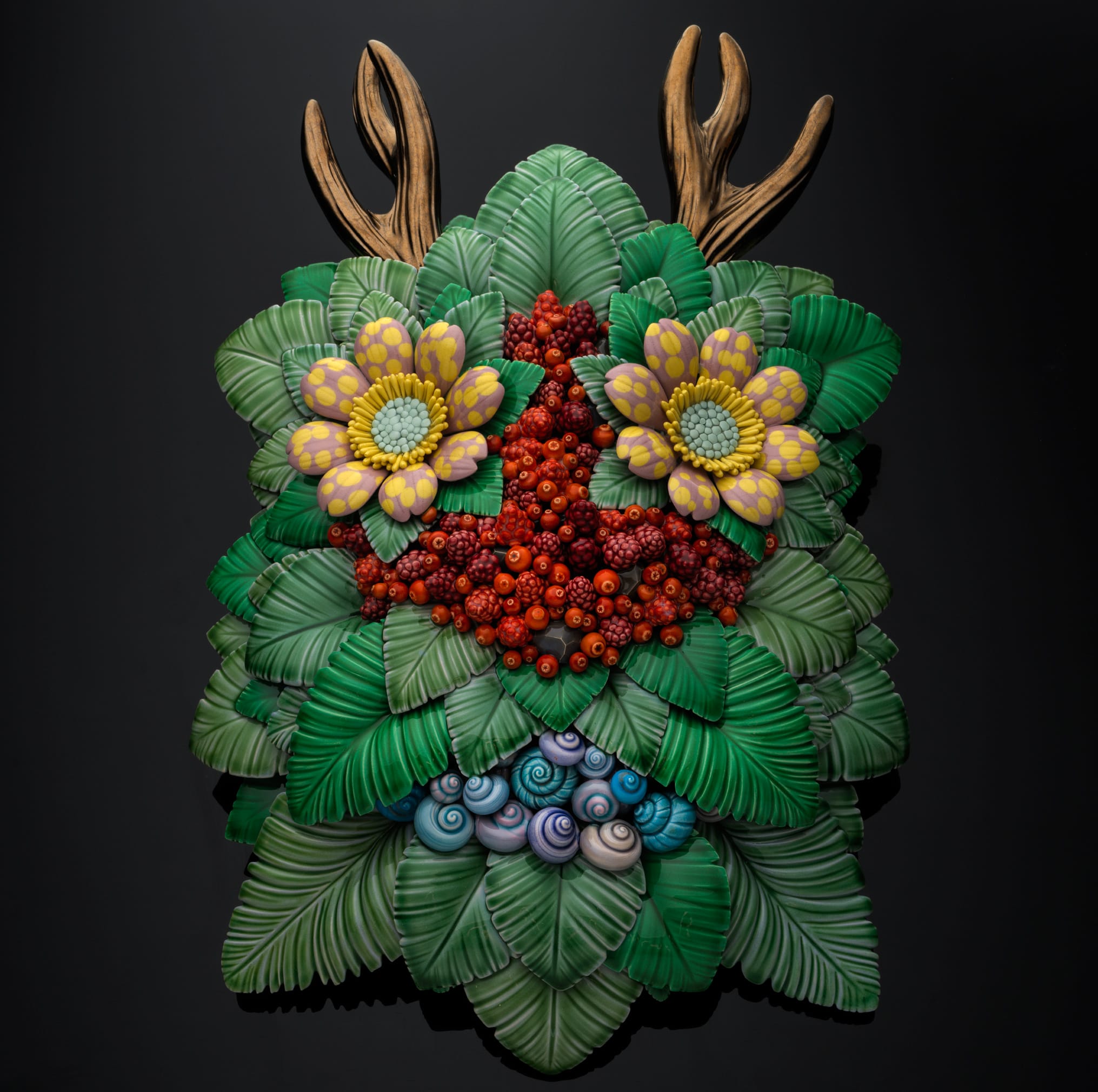 A ceramic sculpture by Male Hartmann Rasmussen of a "troll" face made of green leaves, shells, flowers, berries, and antlers