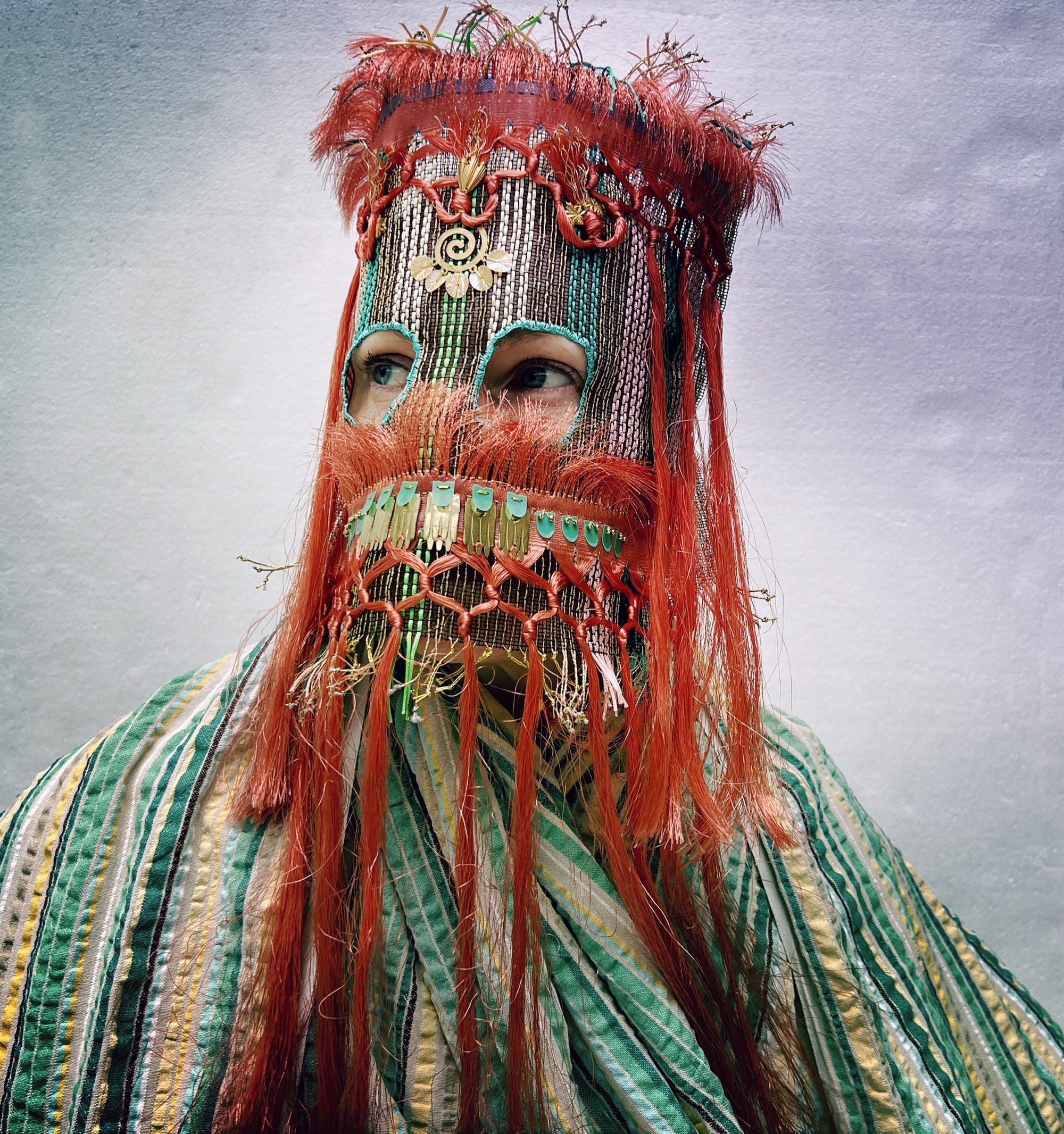 an ornate mask by Magnhild Kennedy with red fringe, blue beads, and other materials and a striped textile covering the body
