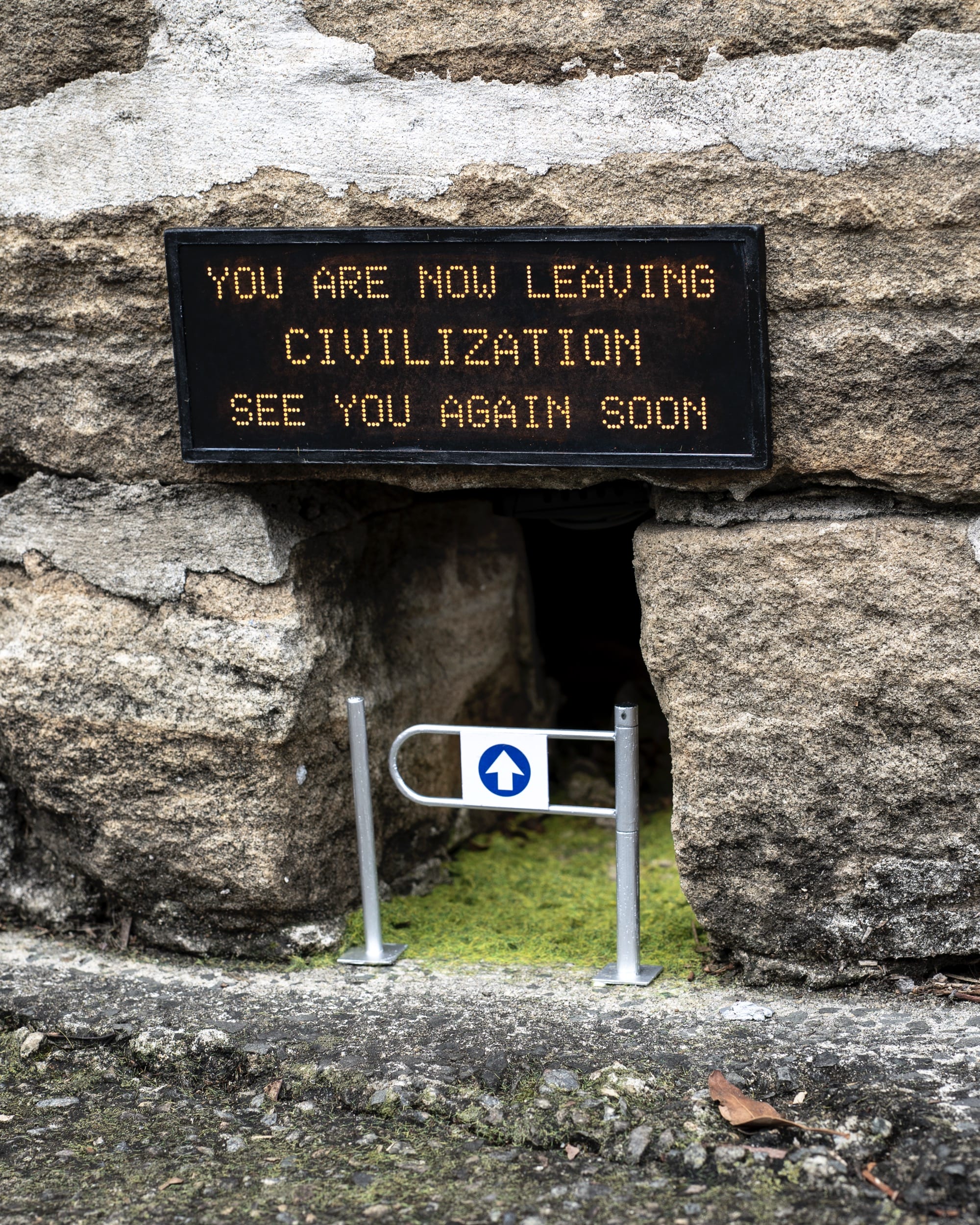 A miniature installation by Michael Pederson of a gap at the base of a stone wall with a gate and a replica digital sign that reads "You are not leaving civilization see you again soon"