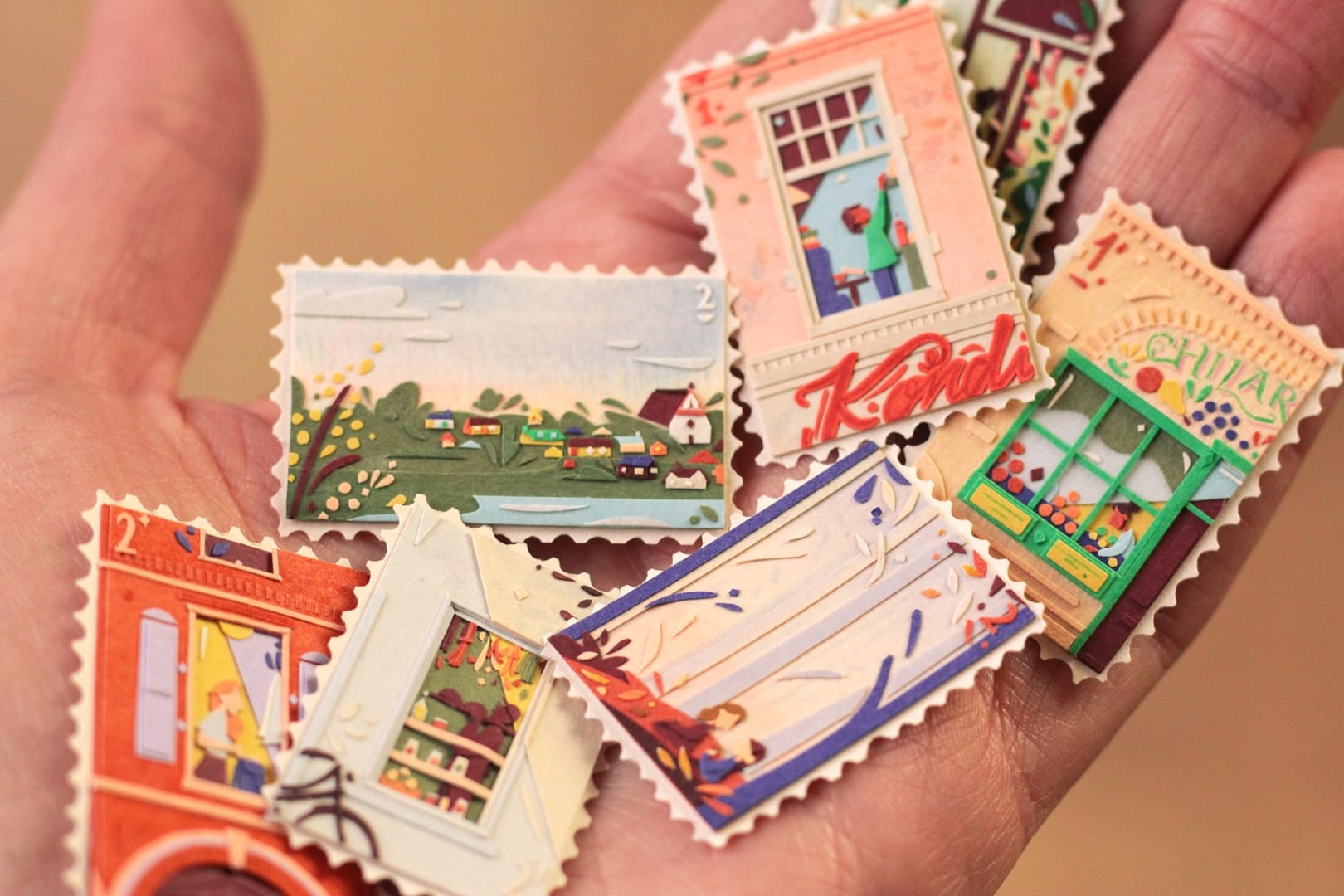 A hand holds a group of paper artworks resembling tiny stamps, featuring landscapes, windows, and trees
