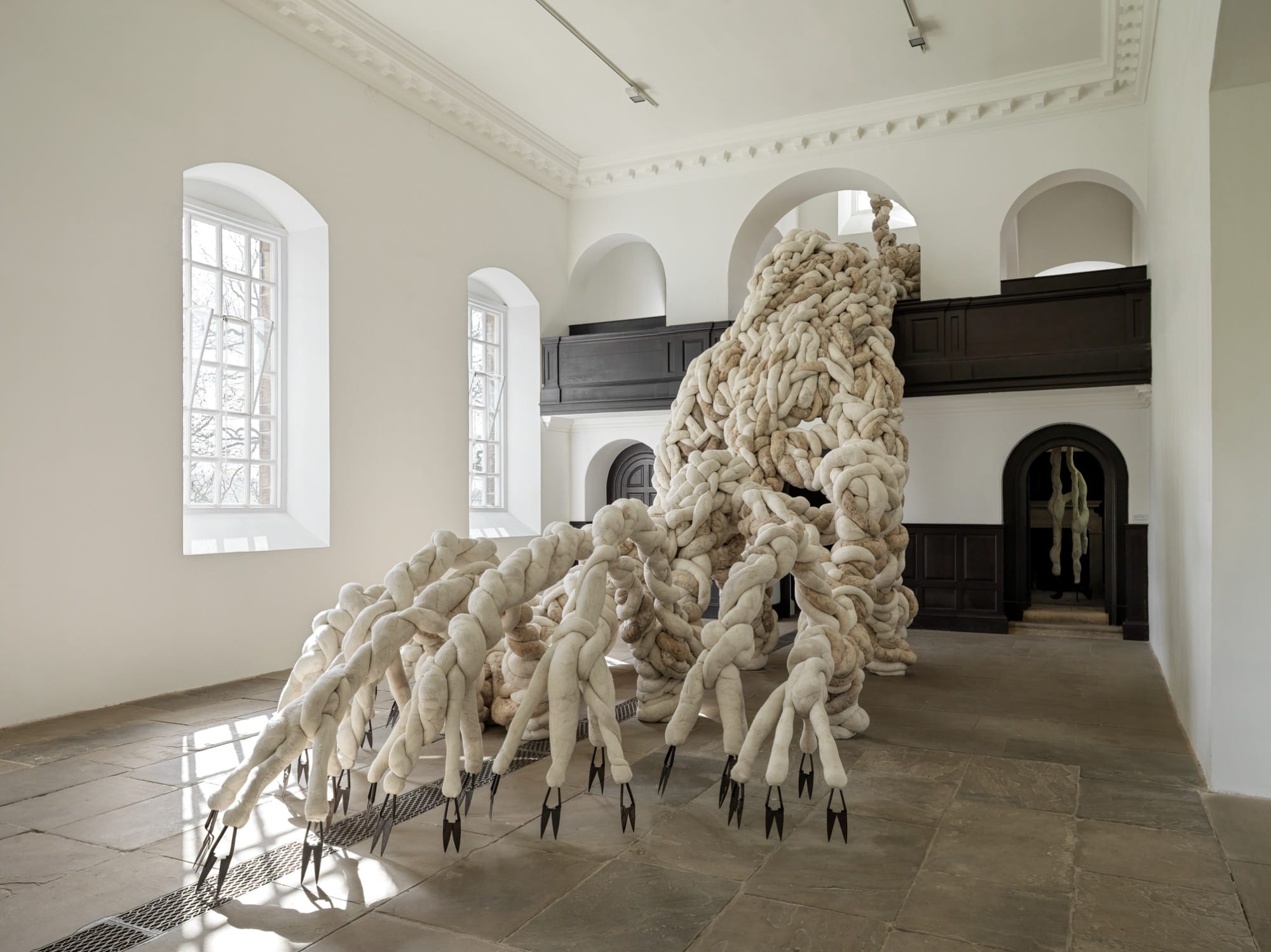 a large-scale installation by Nicola Turner inside of Yorkshire Sculpture Park's 18th-century chapel gallery space of a textile form that appears to be crawling or expanding across the room, out of an opening toward the ceiling, with sharp clippers and scythes for "feet"