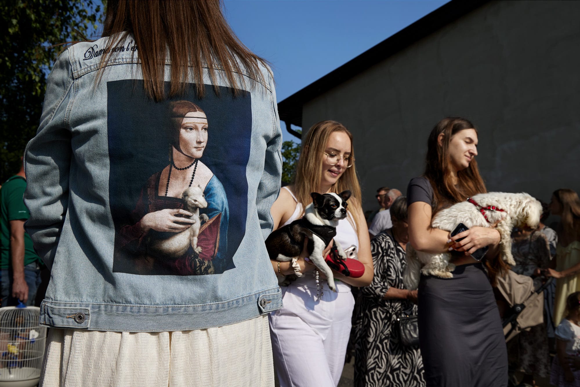 A candid street photograph by Janusz Jurek of women carrying dogs, with a denim jacket with a Leonardo Da Vinci painting of a woman holding an ermine