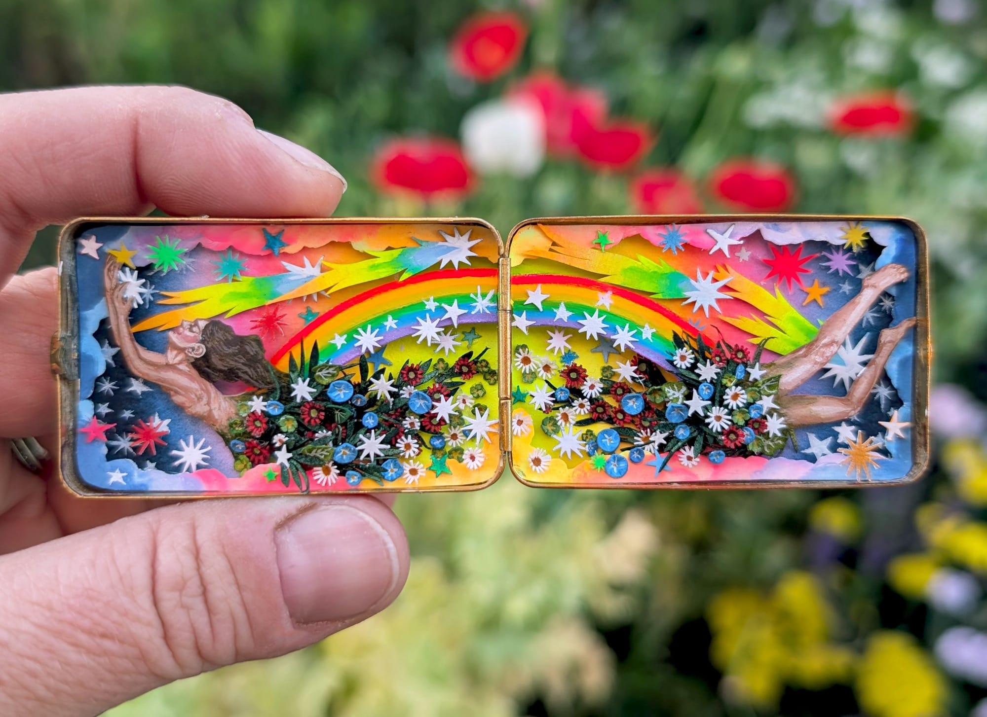a found cosmetic compact filled with a tiny watercolor diorama of a woman soaring through a rainbow-filled sky with flowers for the center of her body