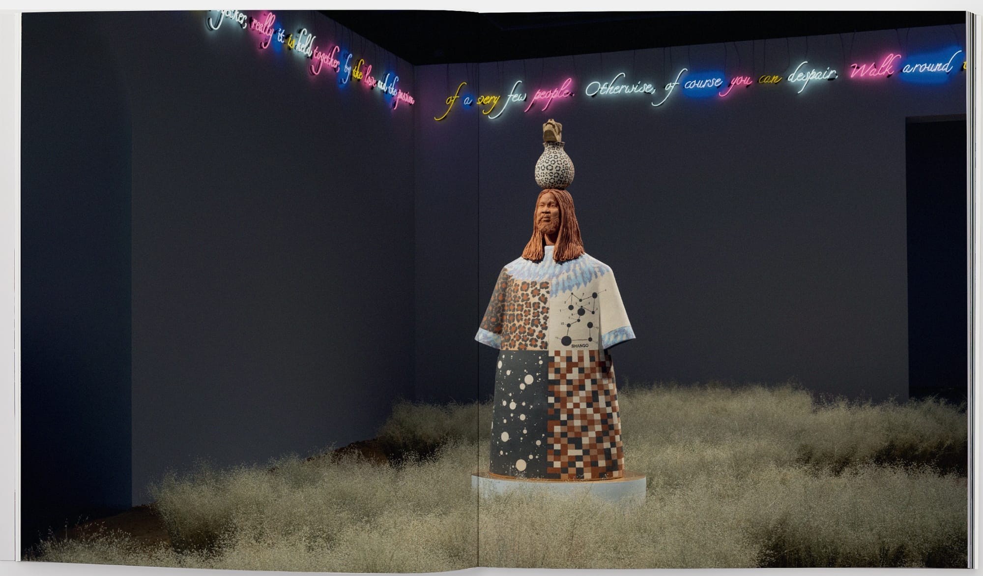 an open book spread with a grassy installation surrounding a figurative sculpture with neon text around the room